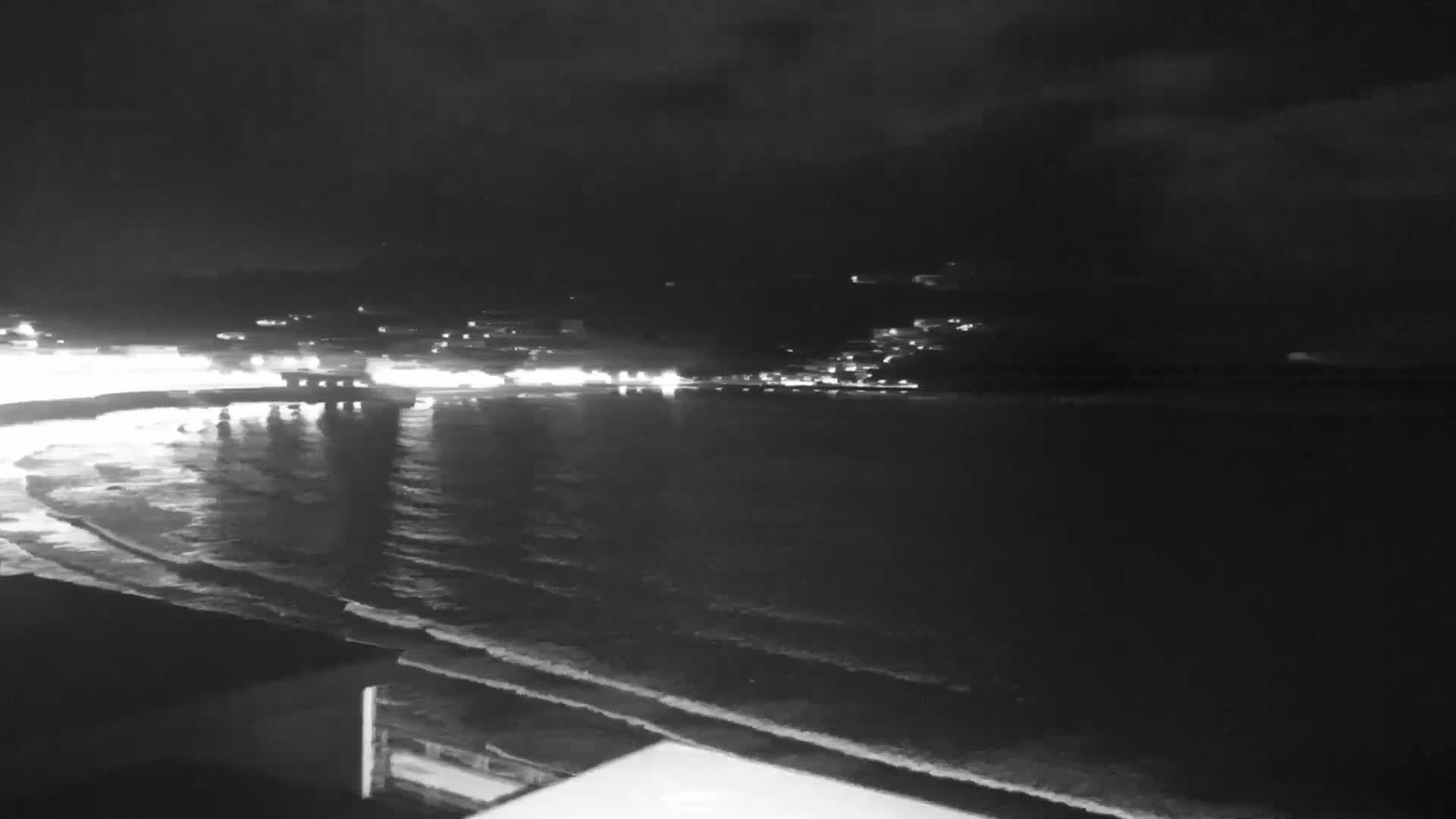 A nighttime view of a coastline shows gently lapping waves on a beach with lights from a town visible in the distance under a dark, cloudy sky.