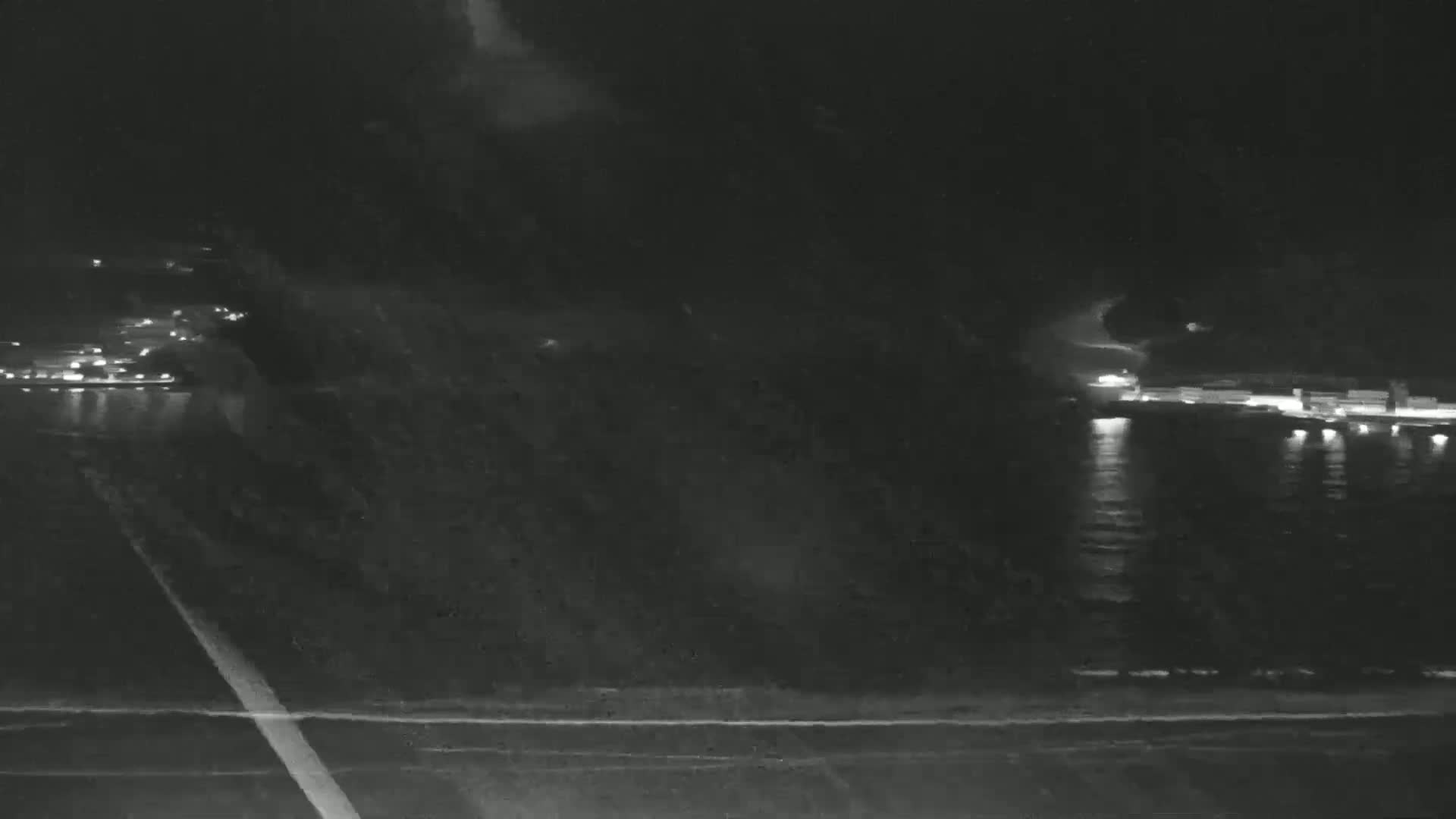 A nighttime, low-light view of a roadway near a body of water with lights visible in the distance on the opposite shore.