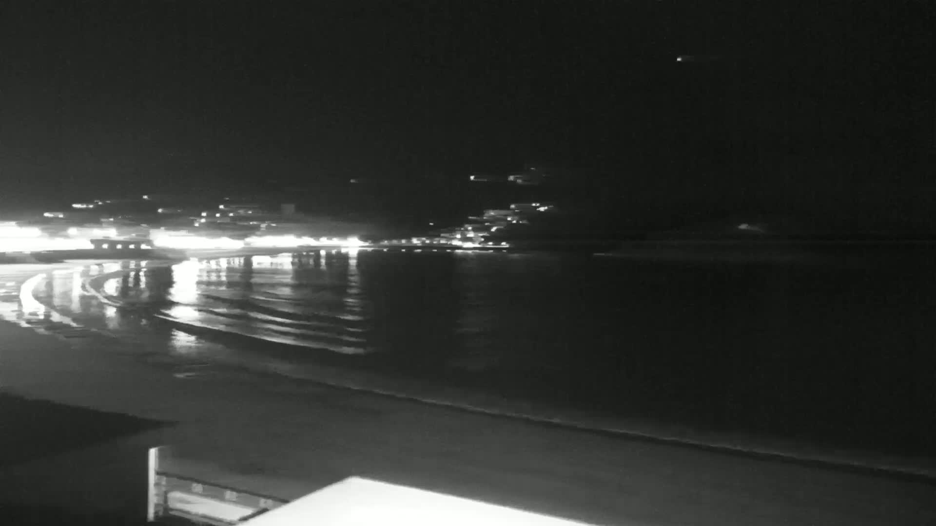 A nighttime view of a calm ocean with gentle waves lapping a beach, illuminated by distant lights on the shore.
