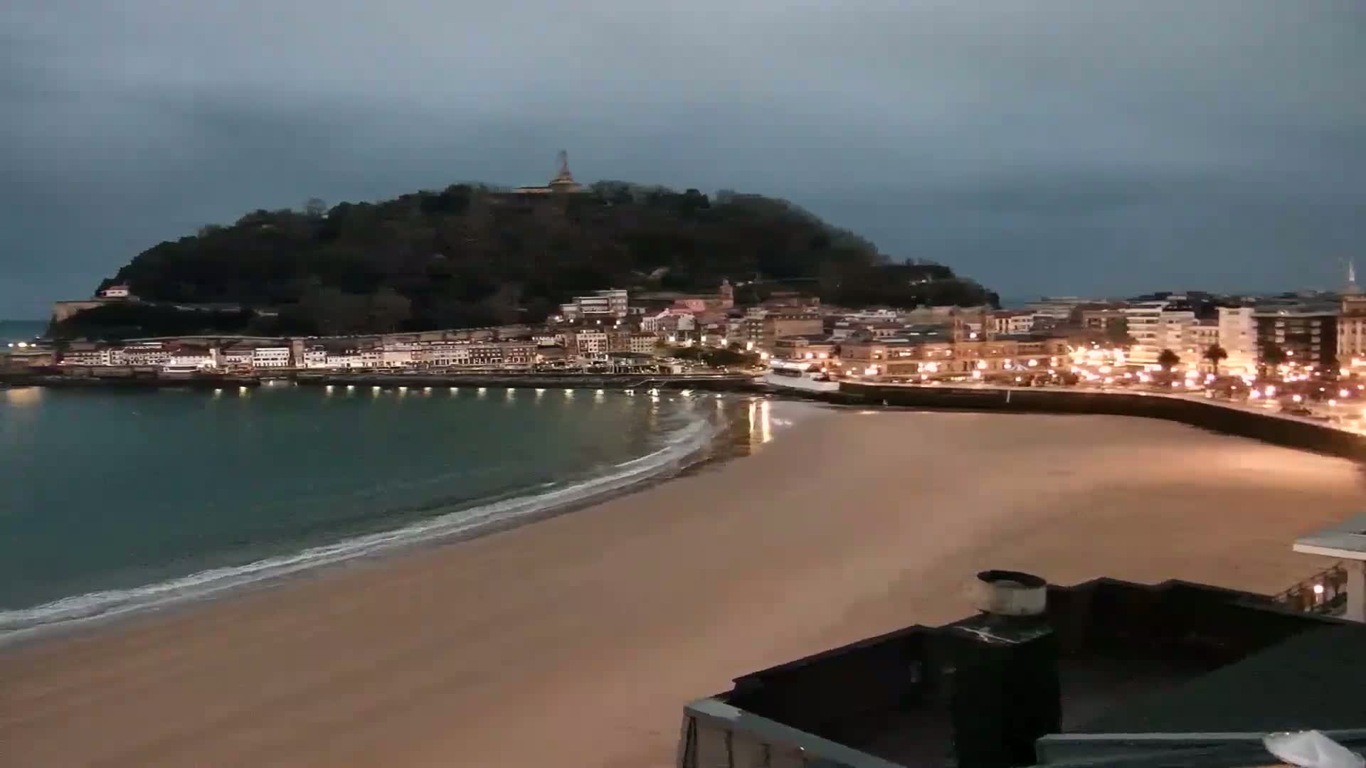 A sandy beach at night, under an overcast sky, curves gently around a calm bay with a town of illuminated buildings nestled at the base of a hill.