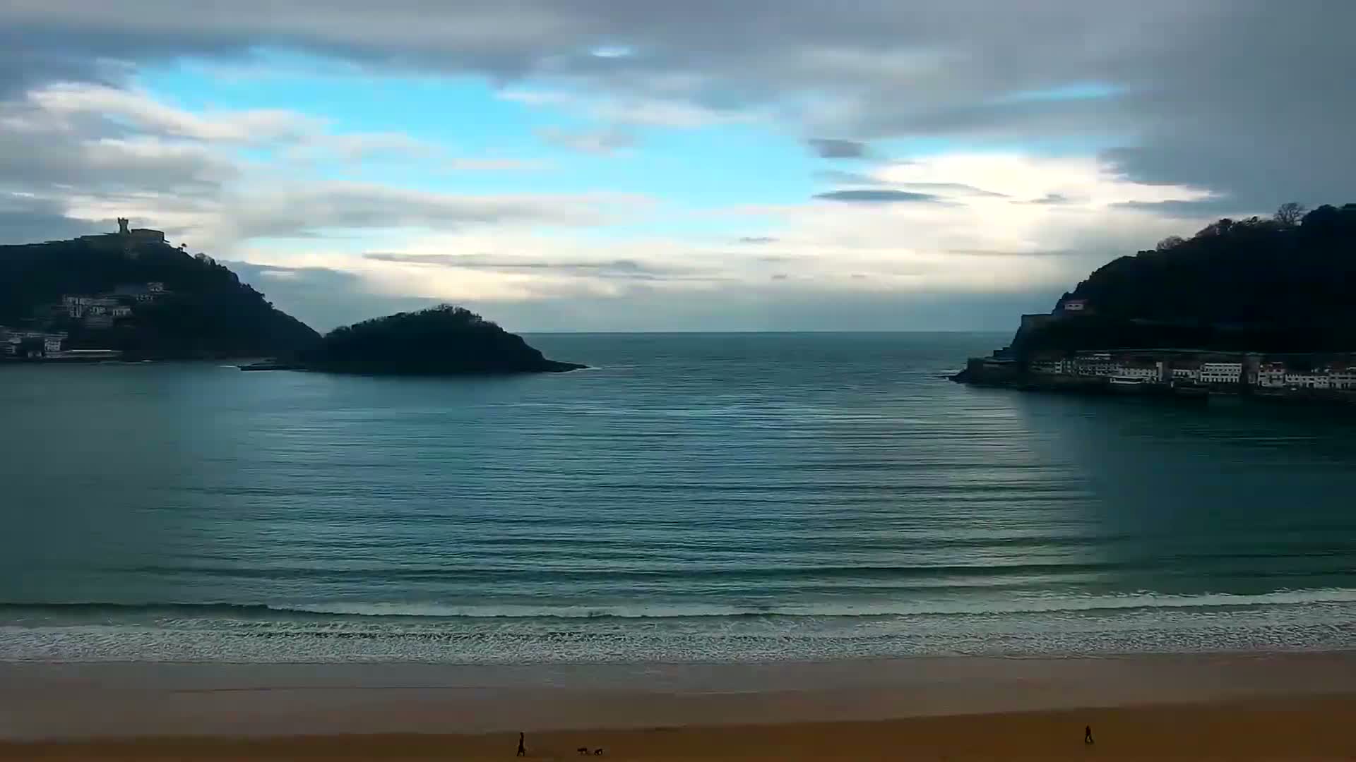 A sandy beach with gentle waves laps the shore, facing a calm bay between two hillsides dotted with buildings, under a mostly cloudy sky.