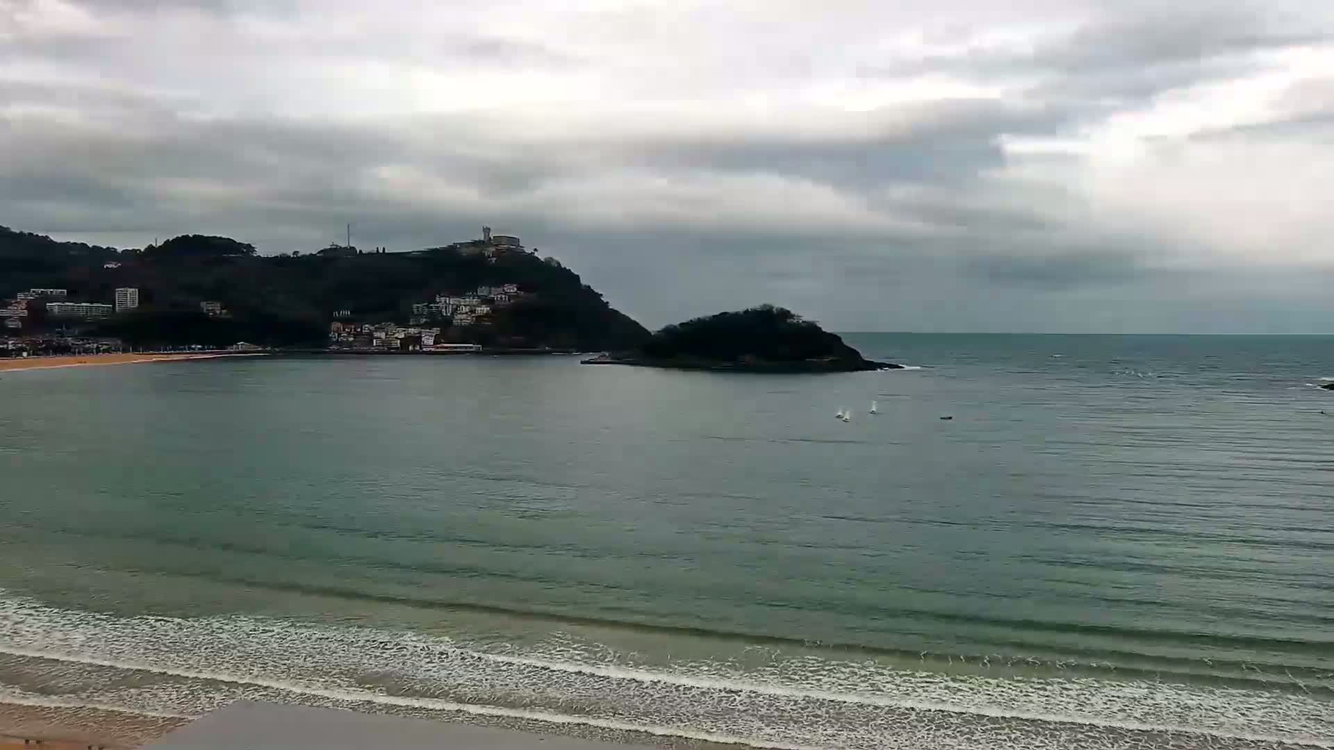 A beach with calm waves under an overcast sky, overlooks a bay with a small island and a hillside town in the distance.