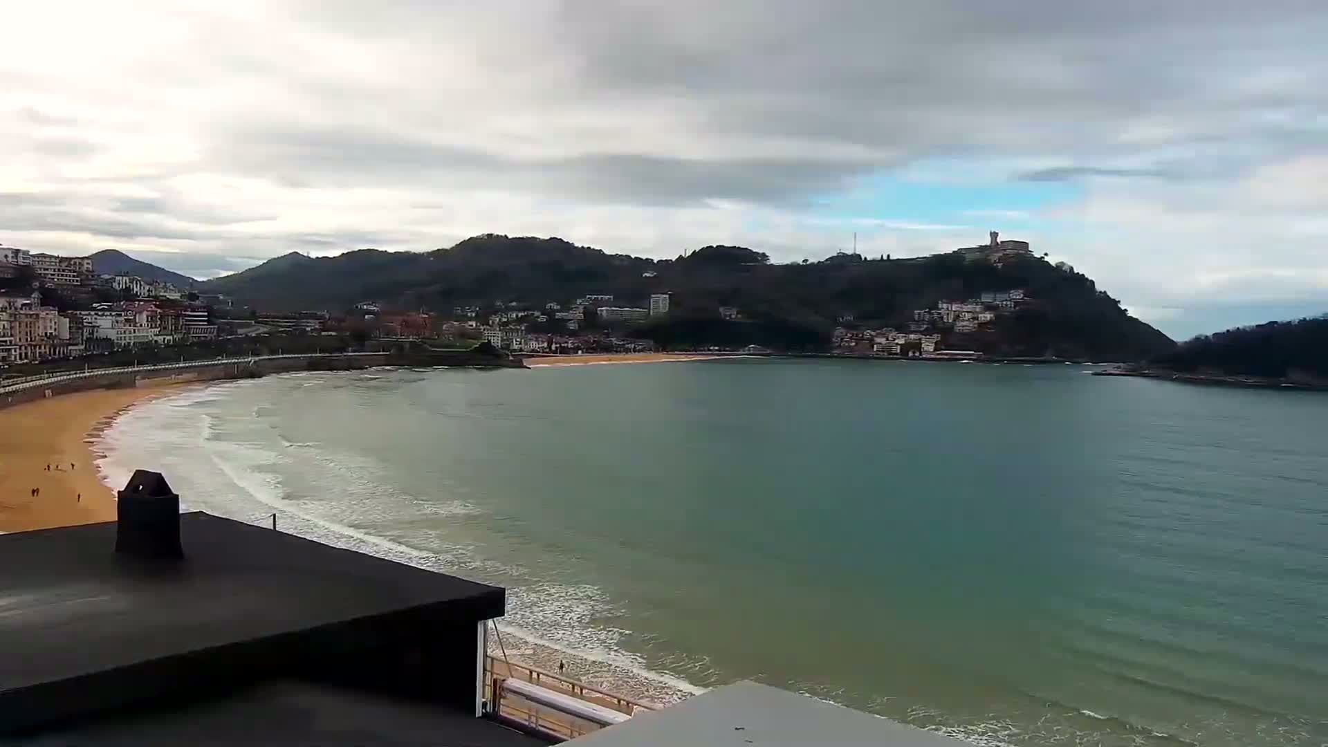 A sandy beach curves around a calm bay, bordered by a town nestled against a hillside under a cloudy sky.