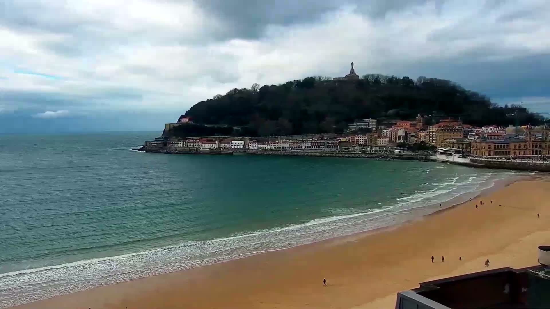 A sandy beach with gentle waves laps the shore below a coastal town nestled at the base of a hill topped with a monument, all under a cloudy sky.