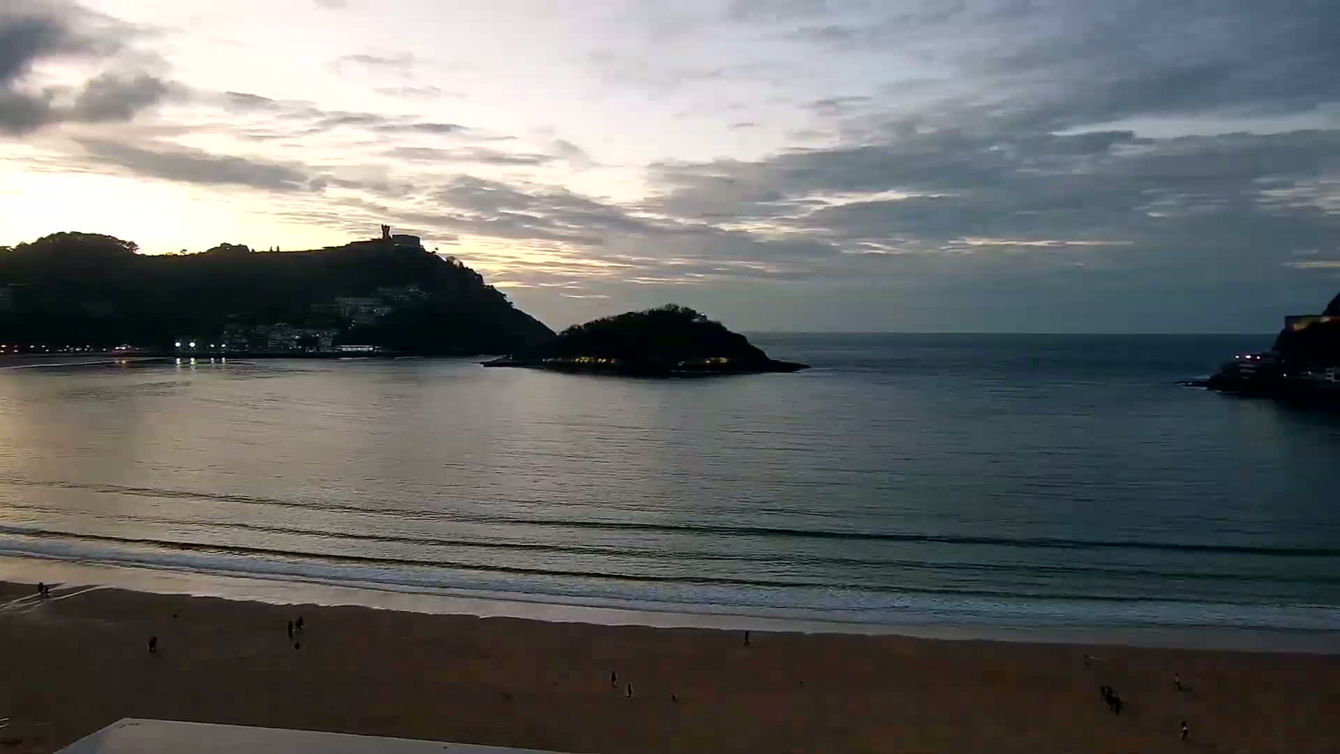 A sandy beach with a few people on it overlooks a calm bay at sunset, with two hills in the distance and a small island separating them.