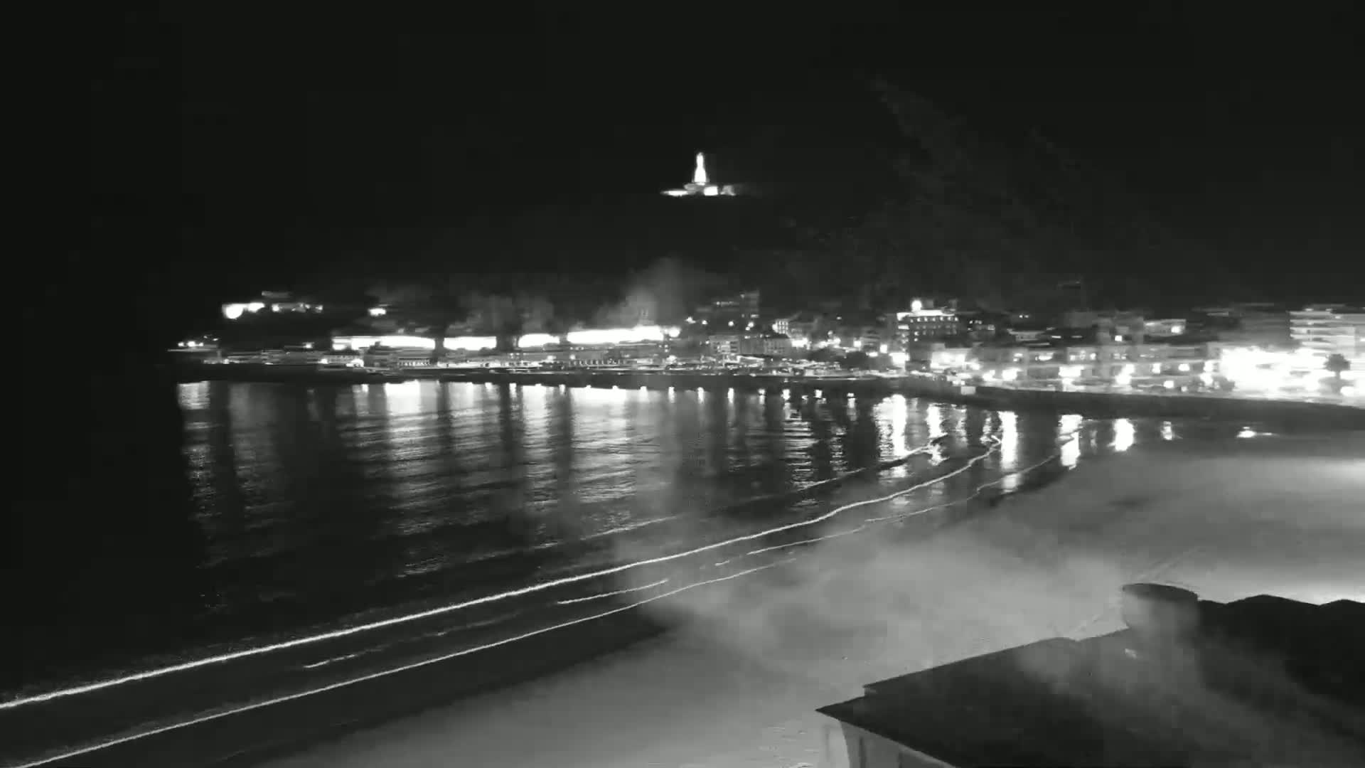 A nighttime view of a calm ocean beach with gently lapping waves, a brightly lit town across the water, and a hilltop structure in the distance.