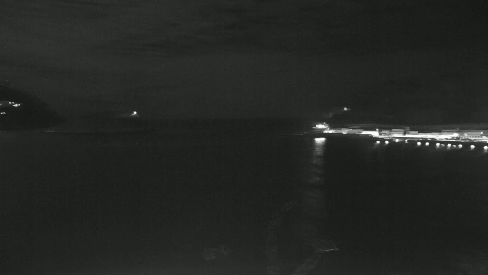A nighttime, low-light view shows a dark body of water with a distant shoreline featuring several illuminated buildings and structures.