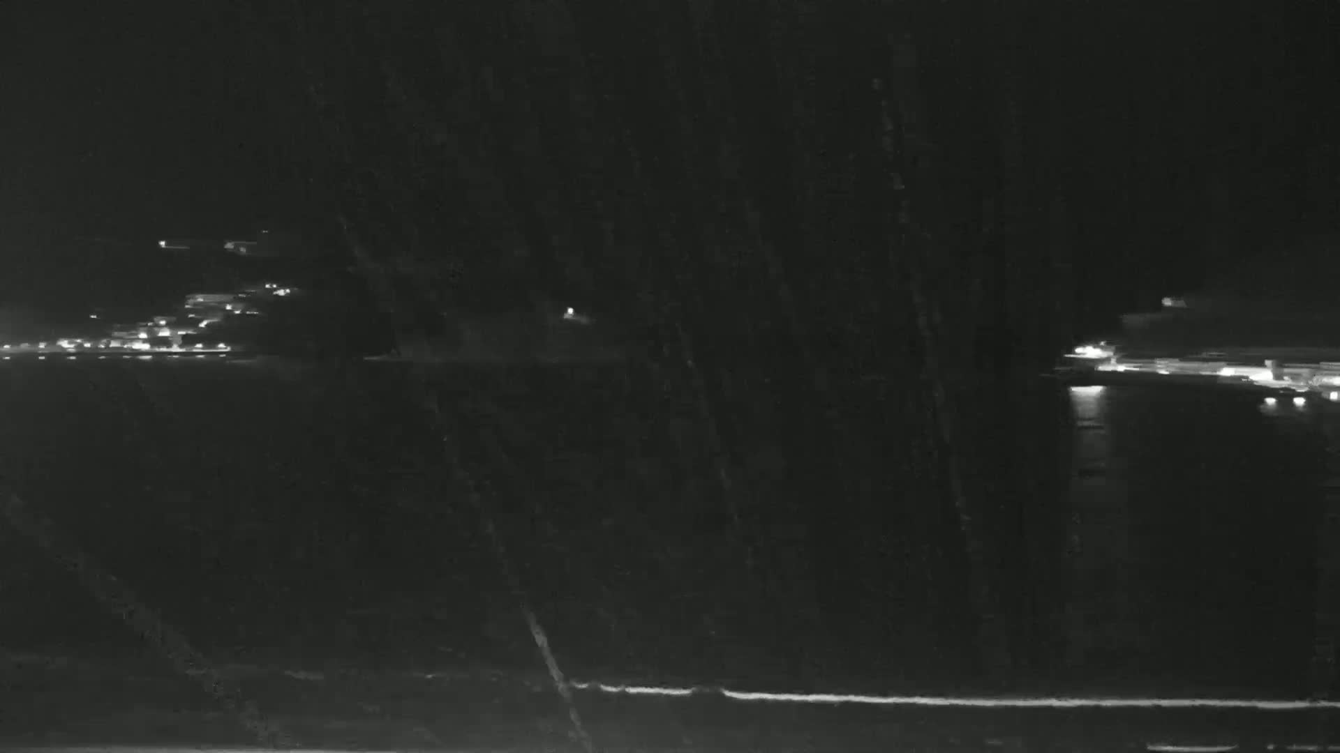 A nighttime long shot shows faint lights across a body of water, with possibly some snow or rain obscuring the view.
