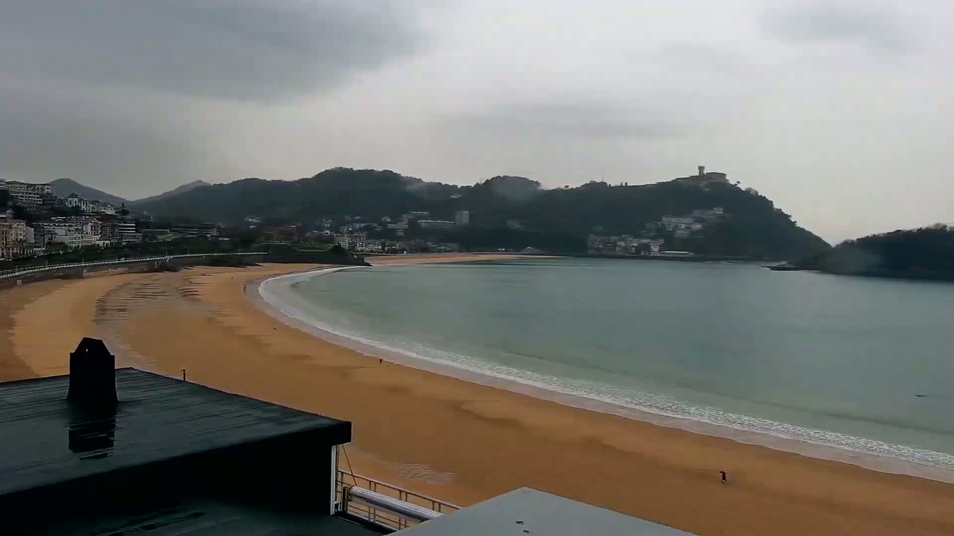 A sandy beach curves around a calm bay under a cloudy, overcast sky, with buildings and hills visible in the distance.