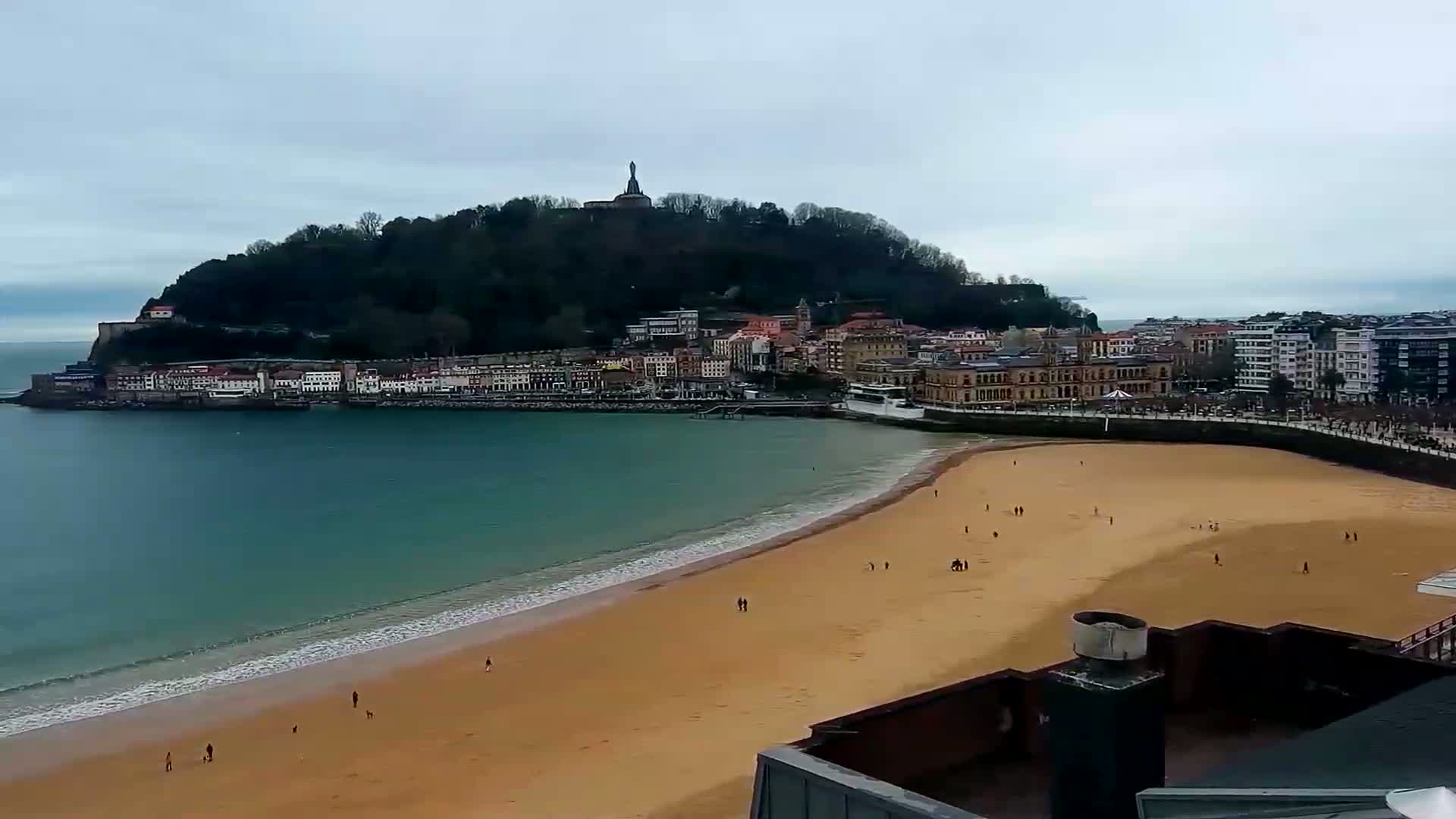 A sandy beach curves around a bay, in front of a town nestled at the base of a tree-covered hill with a statue at its peak, all under an overcast sky.