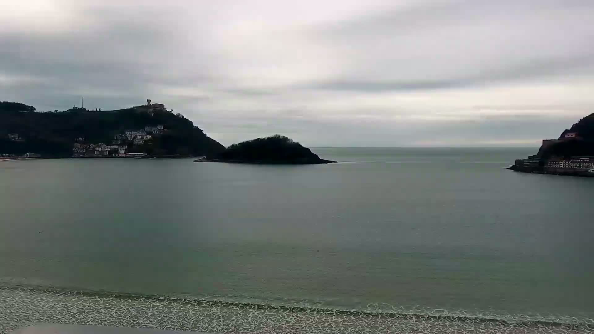 A tranquil bay is viewed from a distance, showcasing a small island in the middle, flanked by hills that are populated with buildings, under a cloudy sky.