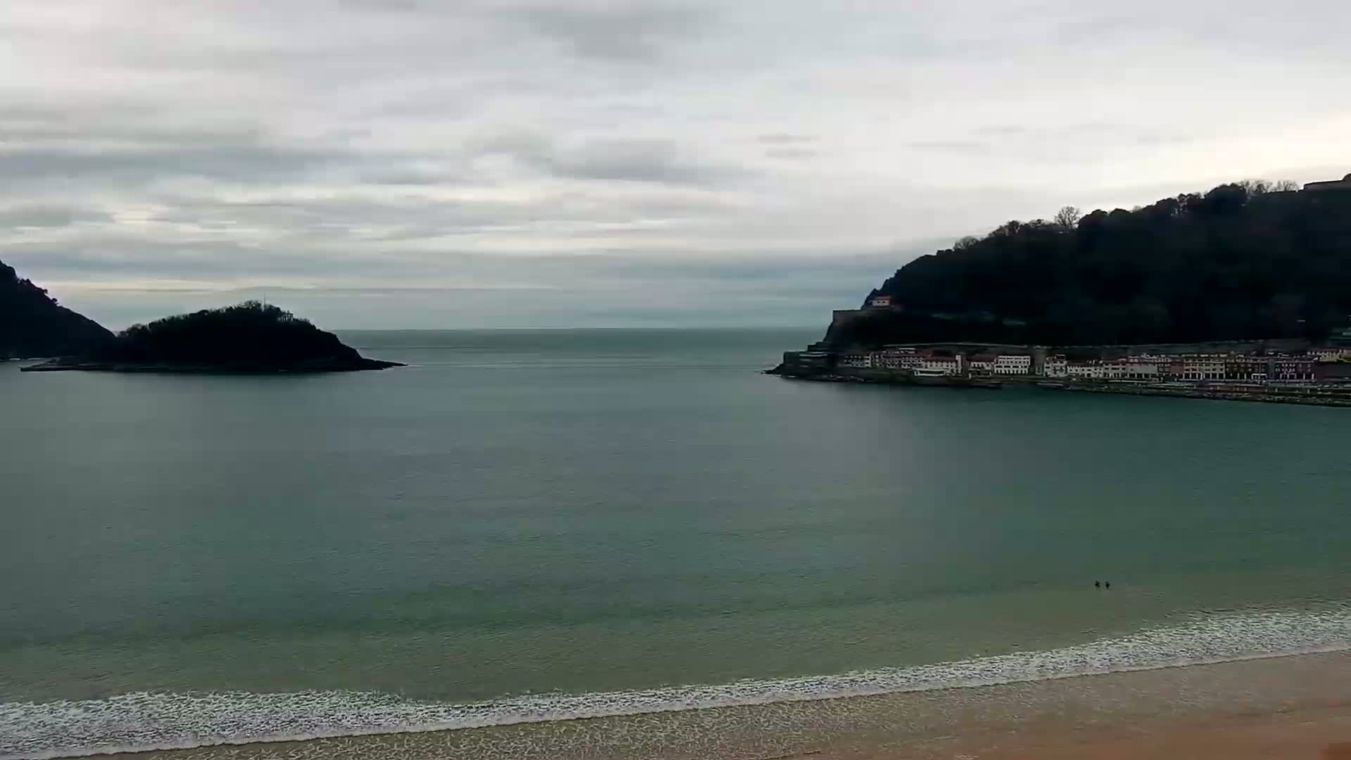 A calm, overcast day reveals a coastal scene with a small island and a hilltop town separated by a bay, with a sandy beach in the foreground.