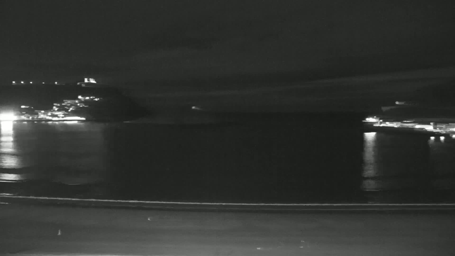 A nighttime view of a dark body of water reflecting the lights of a town on a distant hillside.