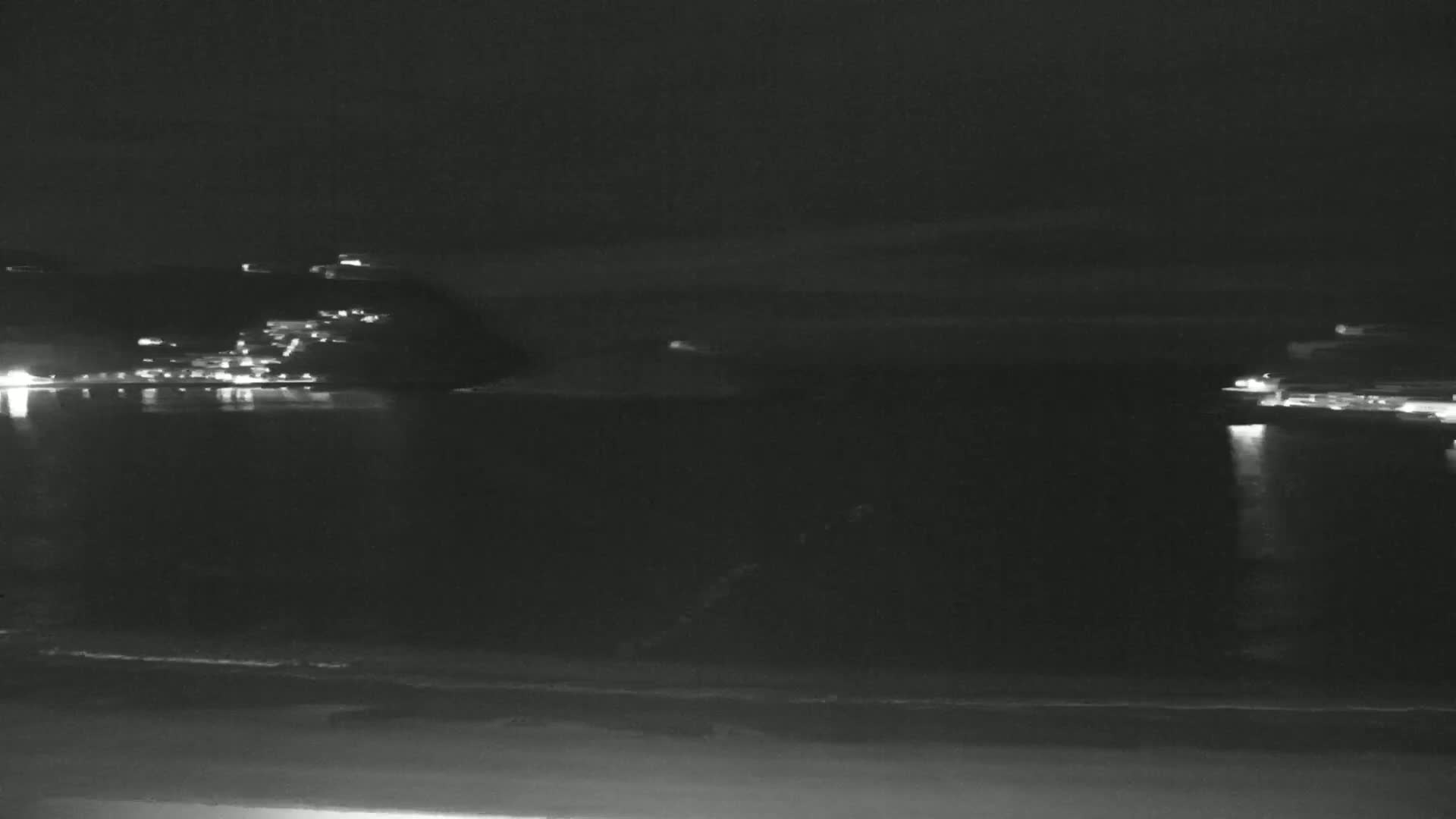 A nighttime coastal scene shows a dark body of water reflecting the lights of buildings on the opposite shore.