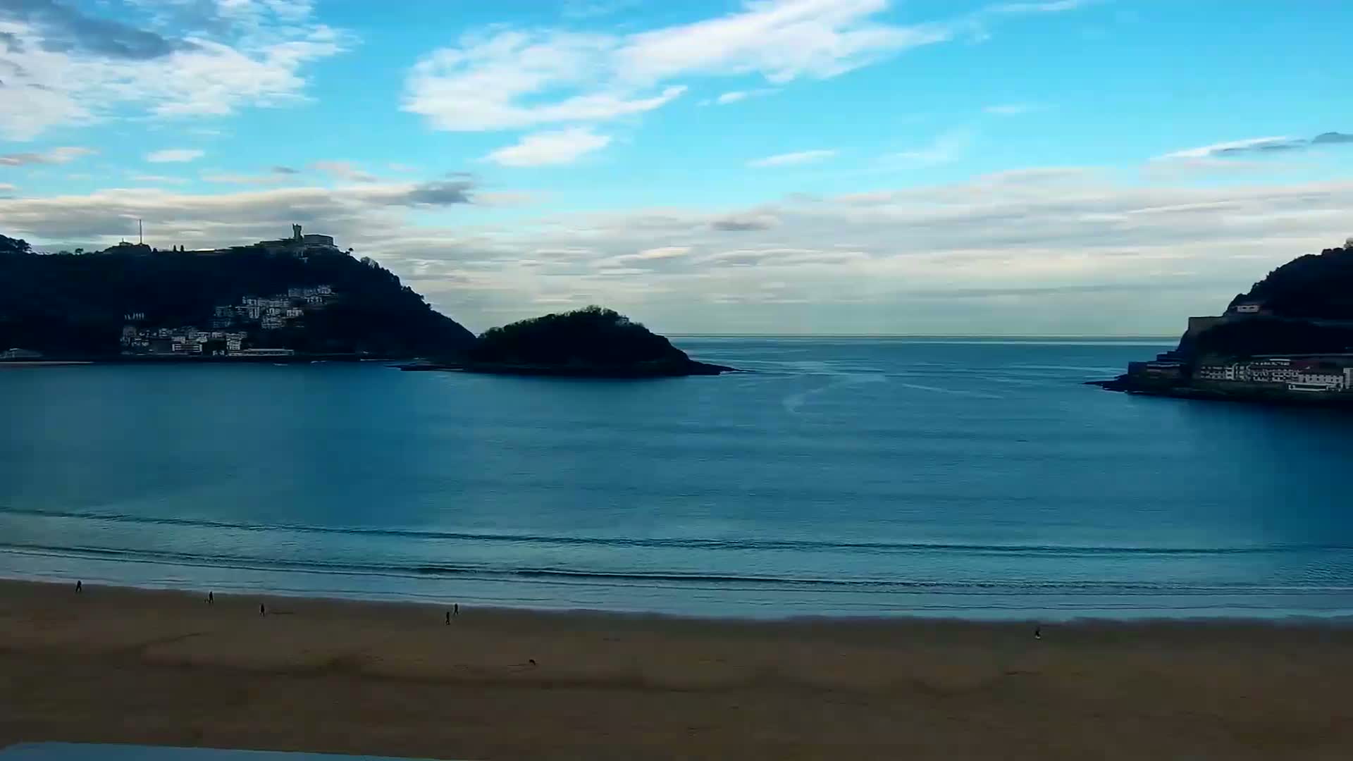 A sandy beach with a few people on it fronts a calm, blue bay with small hills and buildings visible on either side under a mostly clear, blue sky.