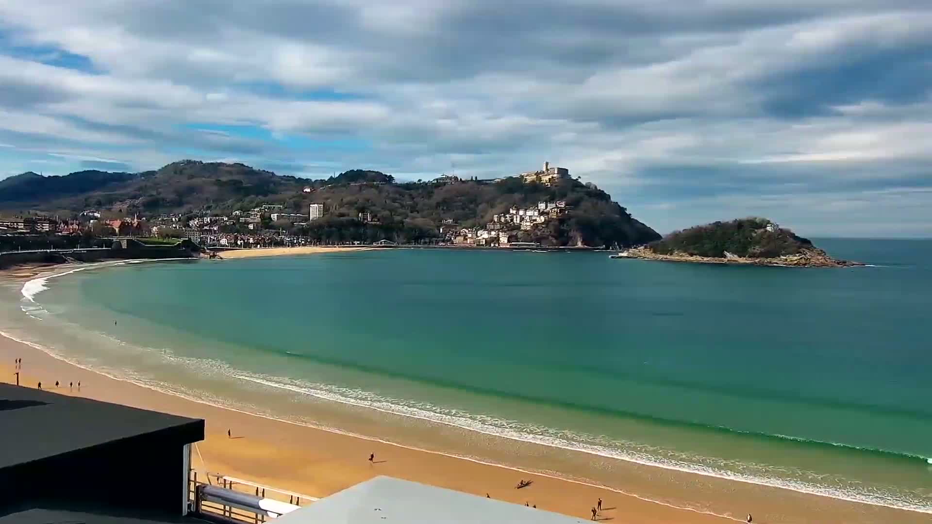 A long sandy beach curves around a bay with teal water, backed by a town nestled against a hill and a small island under a partly cloudy sky.