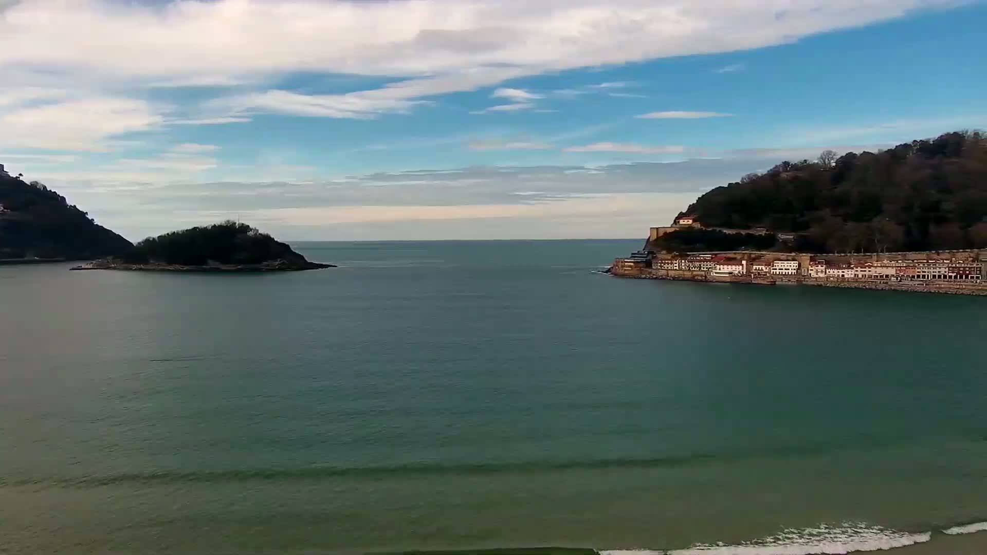 A tranquil ocean scene under a partly cloudy sky shows two hills, one with a small town on it, separated by a calm body of water with small waves on the shore.