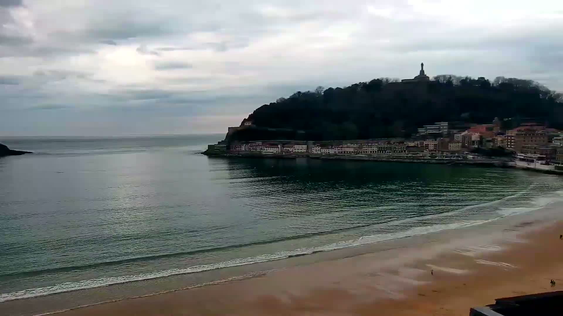 A sandy beach meets a calm, dark-green ocean under a cloudy sky, with a hill topped by a statue and buildings along its shore in the background.