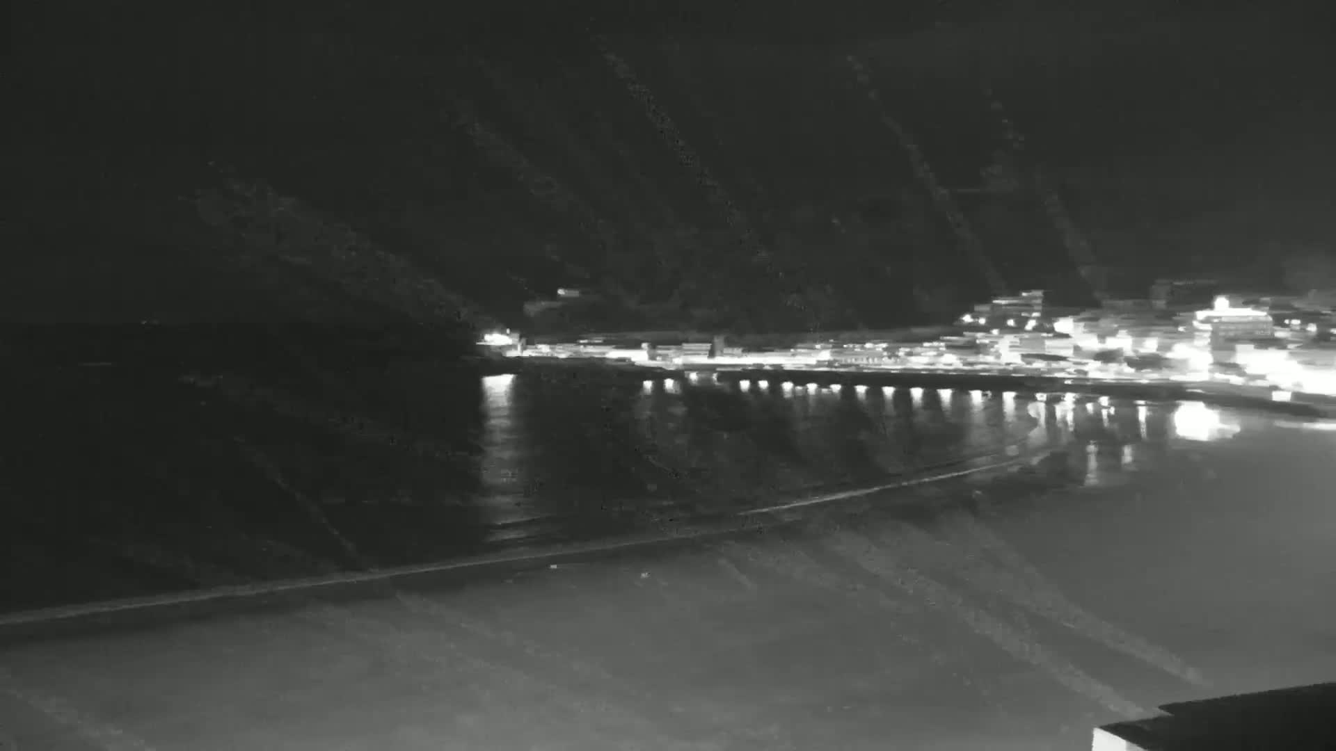 A nighttime, low-light view shows a calm body of water reflecting the lights of a town on the opposite shore.