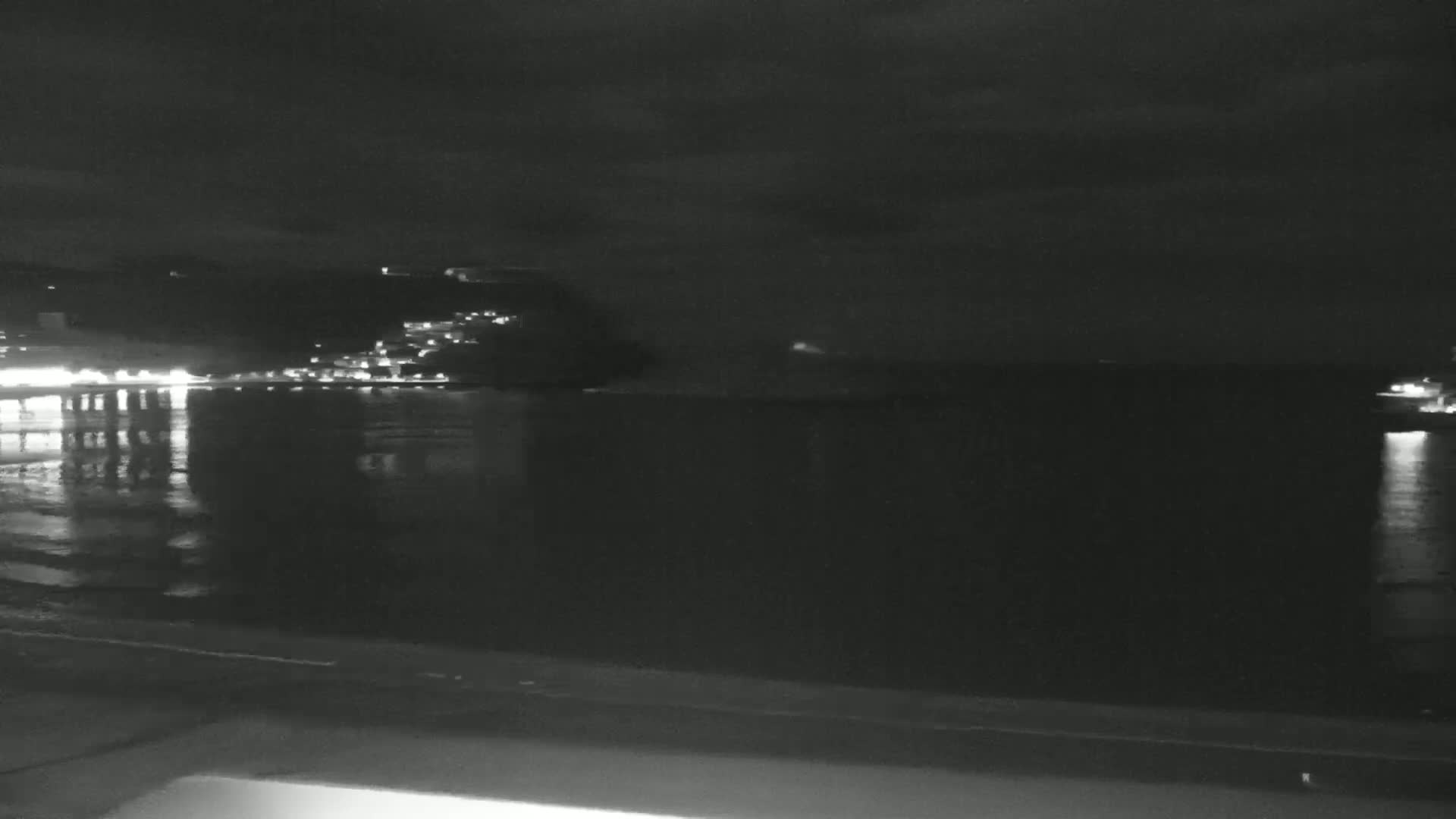 A nighttime view of a calm body of water reflecting the lights of a town on a distant hillside under a dark, cloudy sky.