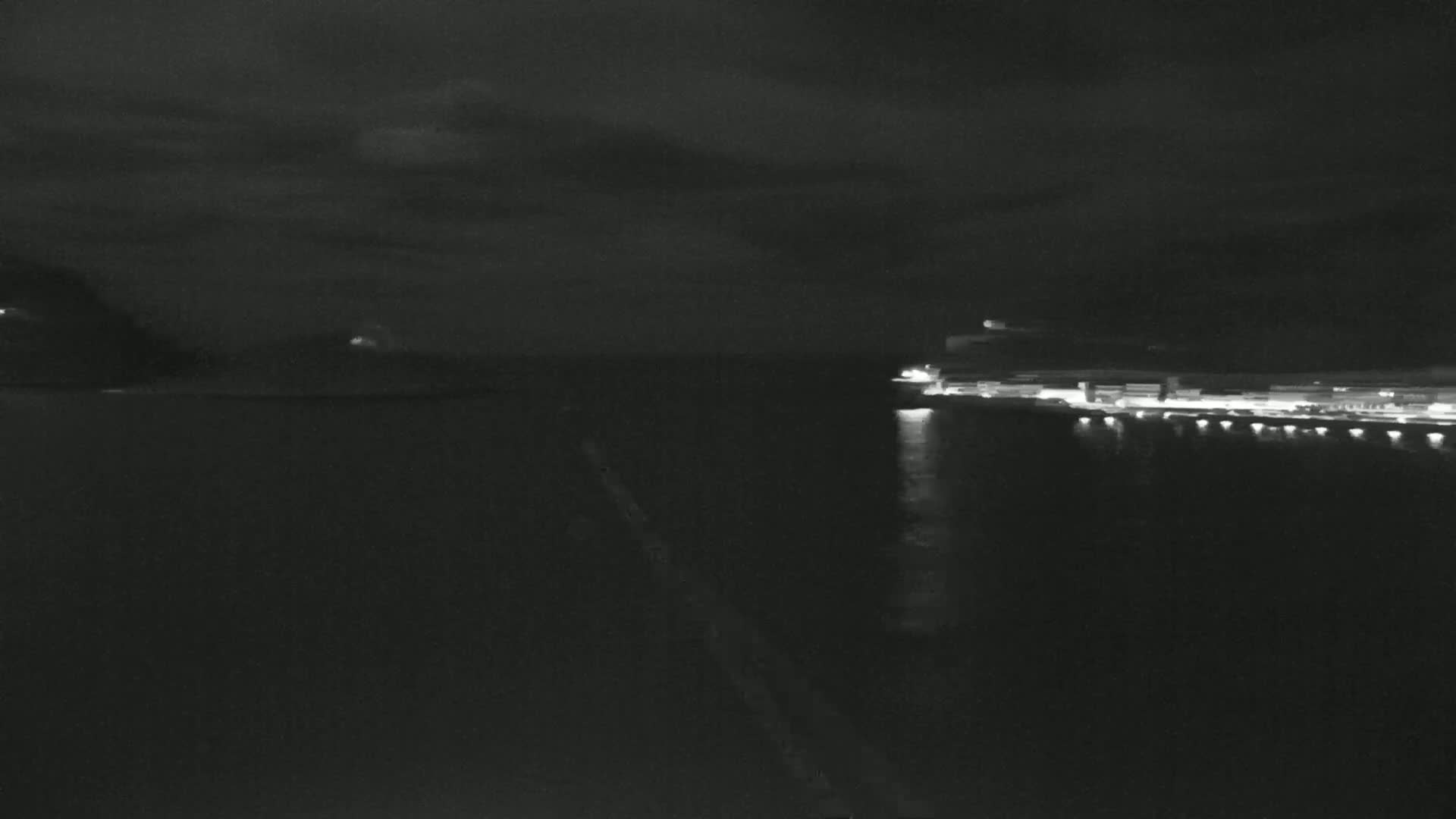 A nighttime view of a dark body of water with a brightly lit shoreline in the distance under a cloudy sky.