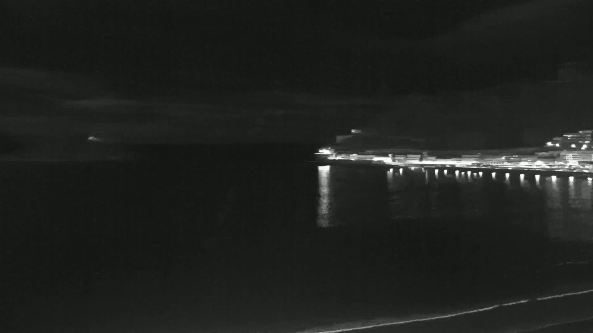 A nighttime view of a city's illuminated waterfront, reflected in a calm body of water under a dark, cloudy sky.