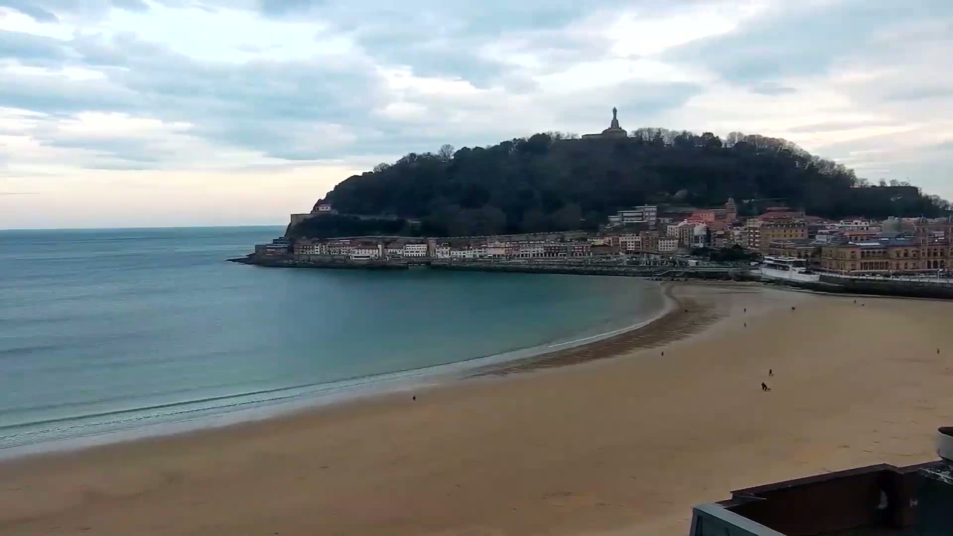 A sandy beach curves around a bay, with a town nestled against a tree-covered hill featuring a statue at its peak, all under a cloudy sky.
