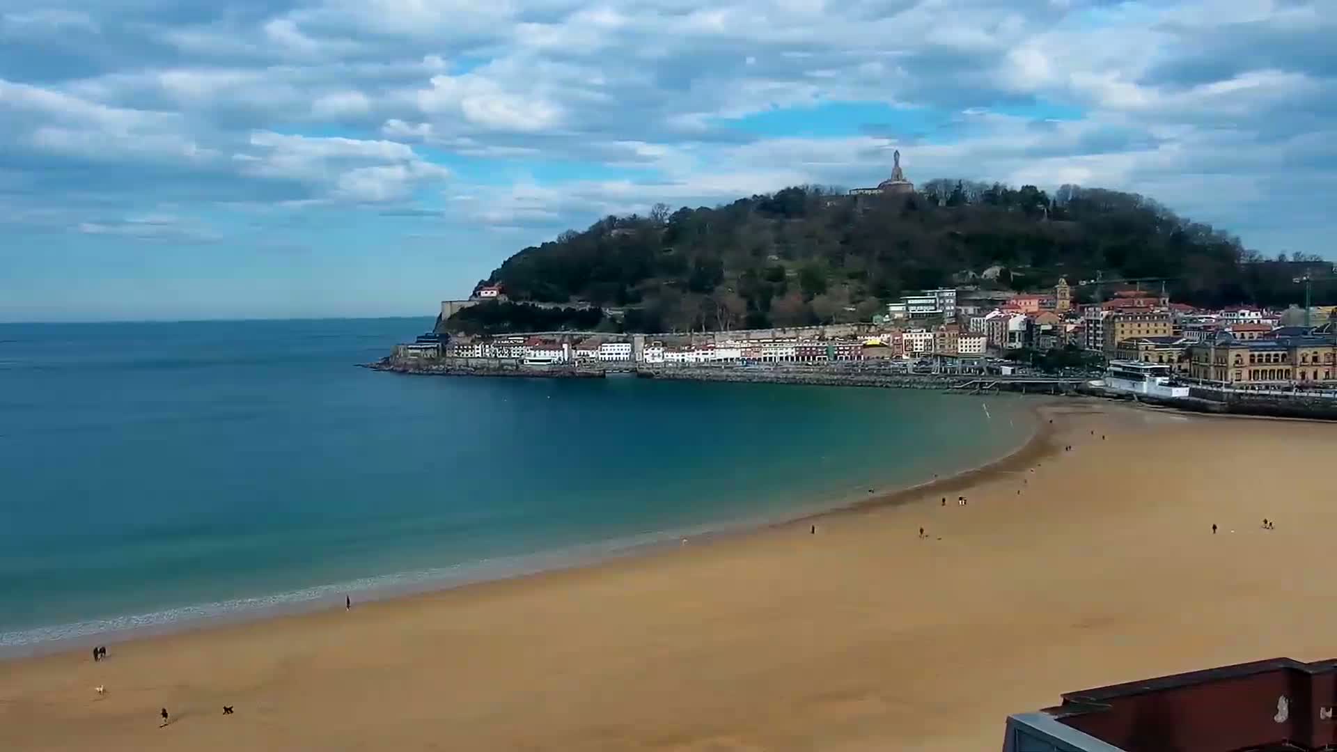 A sandy beach curves along a calm bay, with a town nestled against a hill topped by a monument, under a partly cloudy sky.