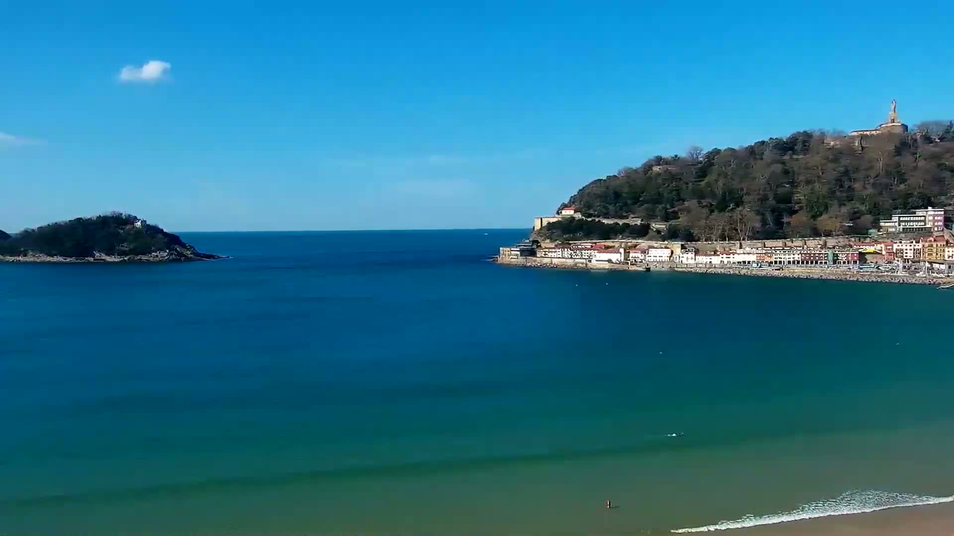 A coastal town nestled beneath a tree-covered hill, with a small island in the distance, under a clear blue sky.