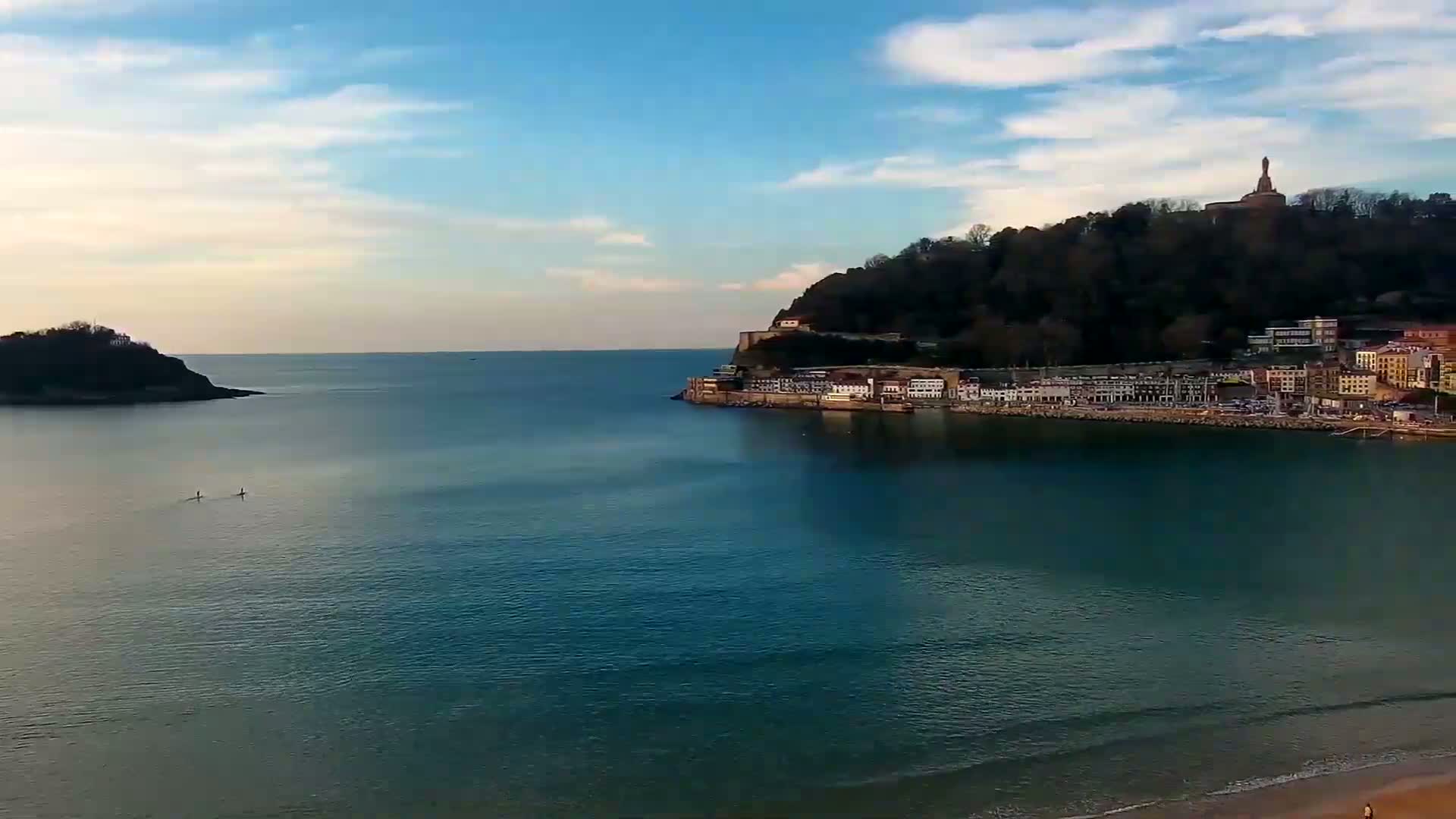 A calm, blue ocean separates a small, dark island from a larger landmass with a town and a hilltop statue, under a partly cloudy sky.
