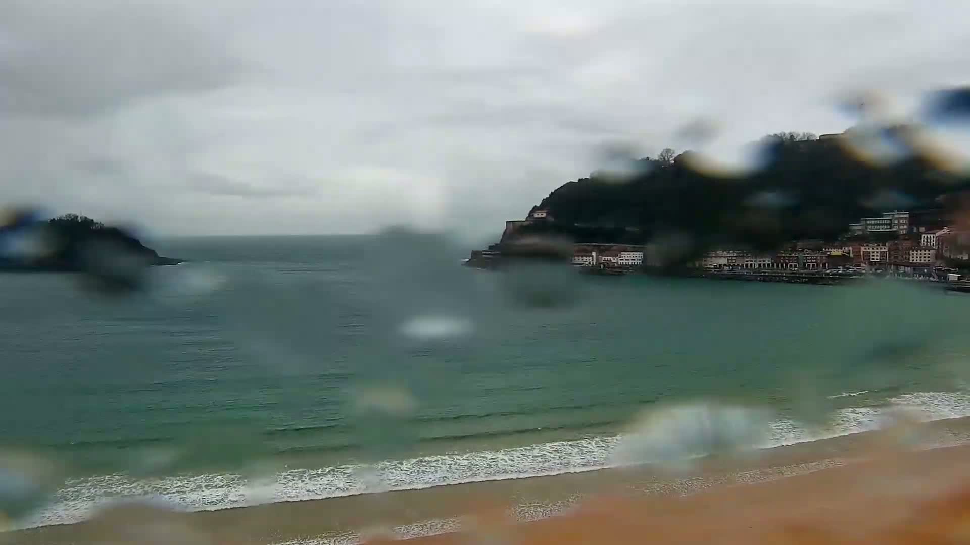 A rain-blurred view shows a sandy beach, waves lapping the shore, a calm bay, and a town built into a hillside on a cloudy day.