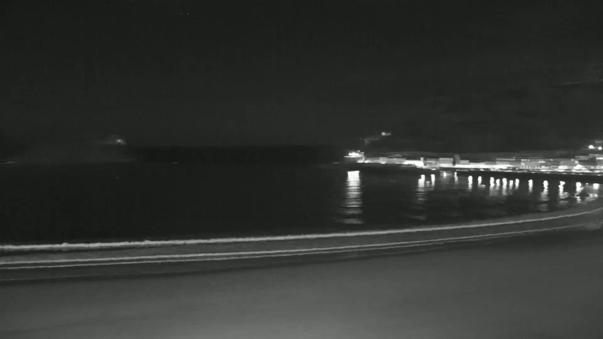 A nighttime view of a dark ocean with small waves lapping a shoreline, lit by the lights of a distant town across the water.