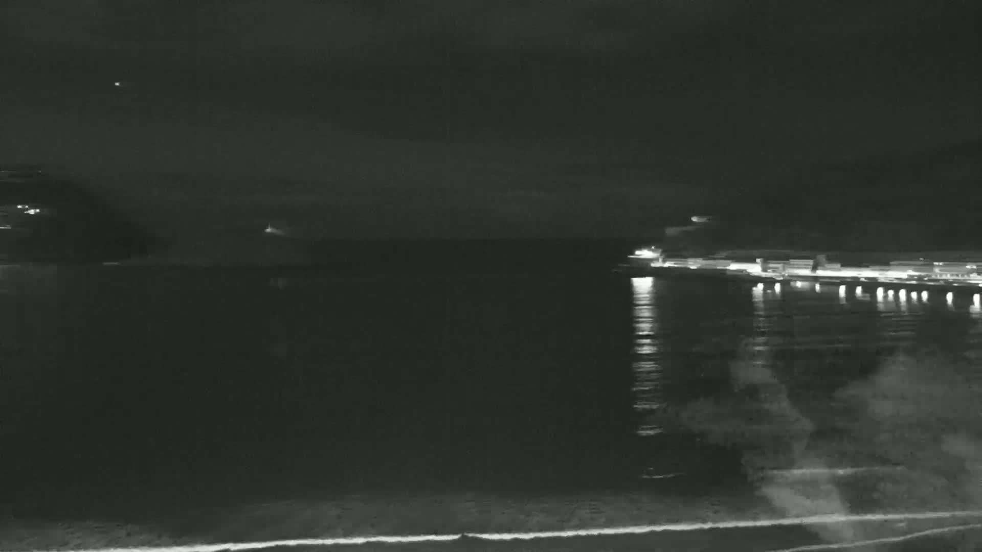 A nighttime view of a calm body of water with a distant shoreline showing lights, under a dark, cloudy sky.