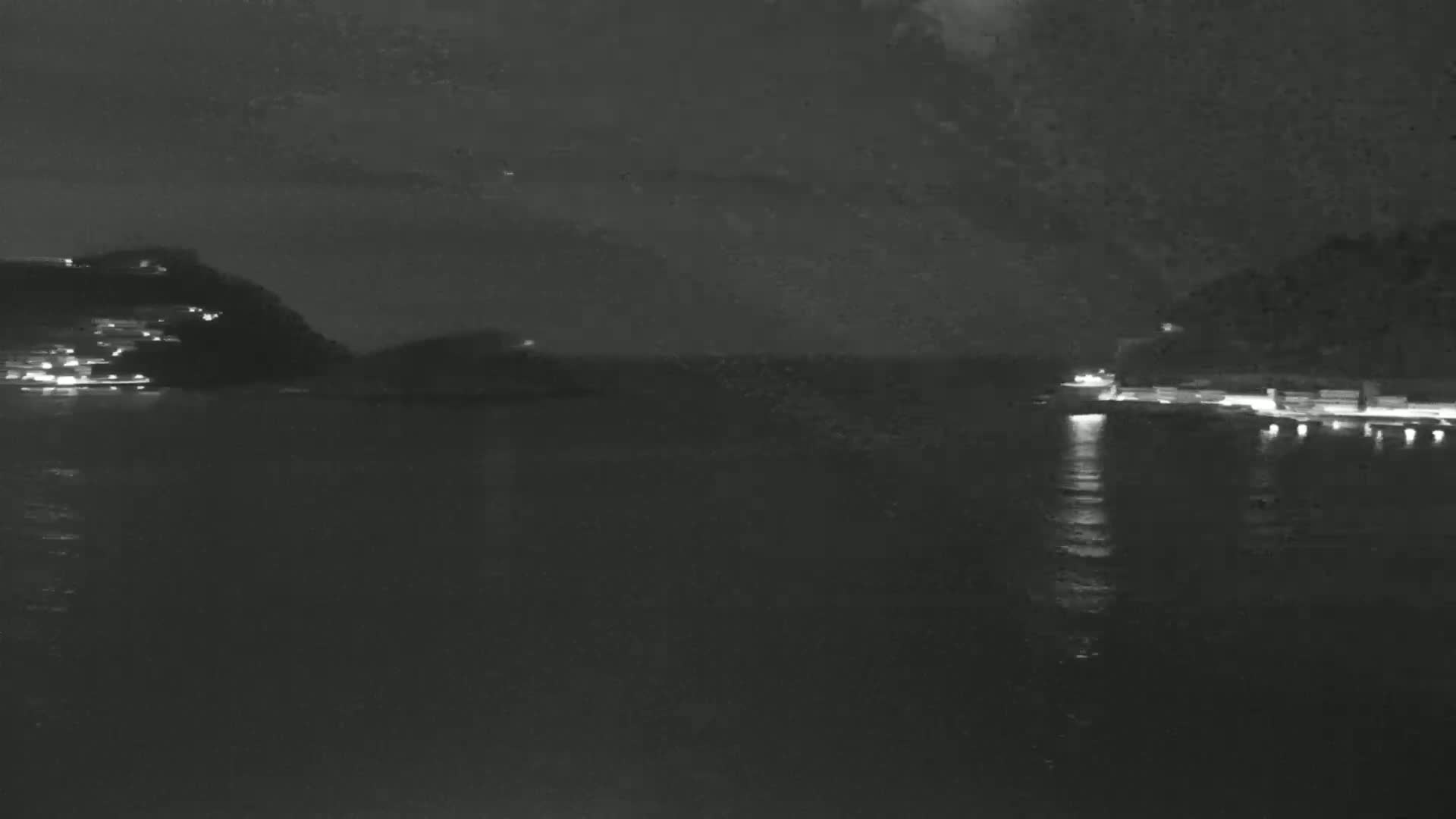 A nighttime view of a dark body of water with dimly lit hillsides on either side, and a few lights visible on the shoreline.