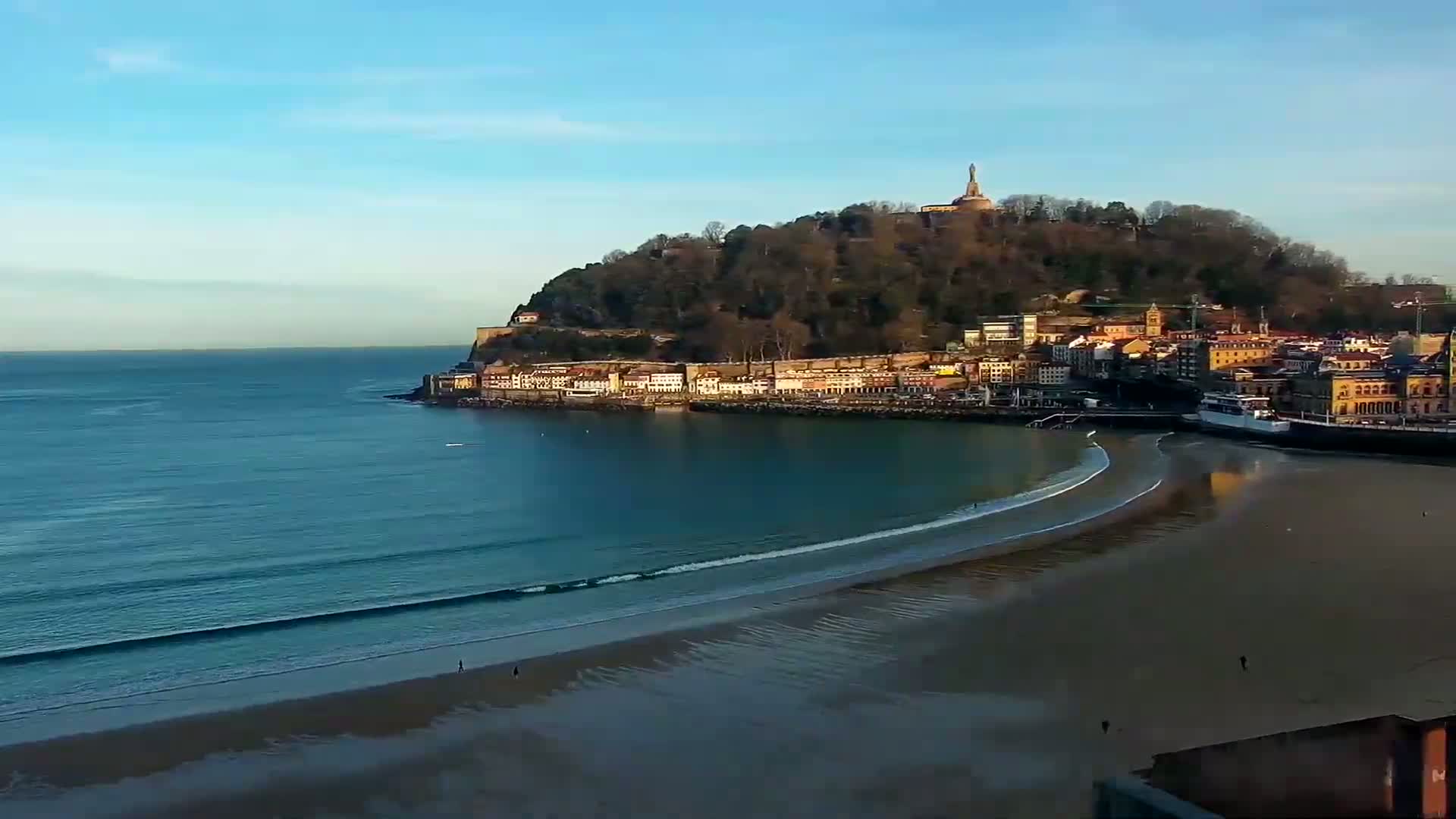 A sandy beach curves gently around a calm bay, next to a town nestled against a tree-covered hill topped with a monument, all under a clear blue sky.