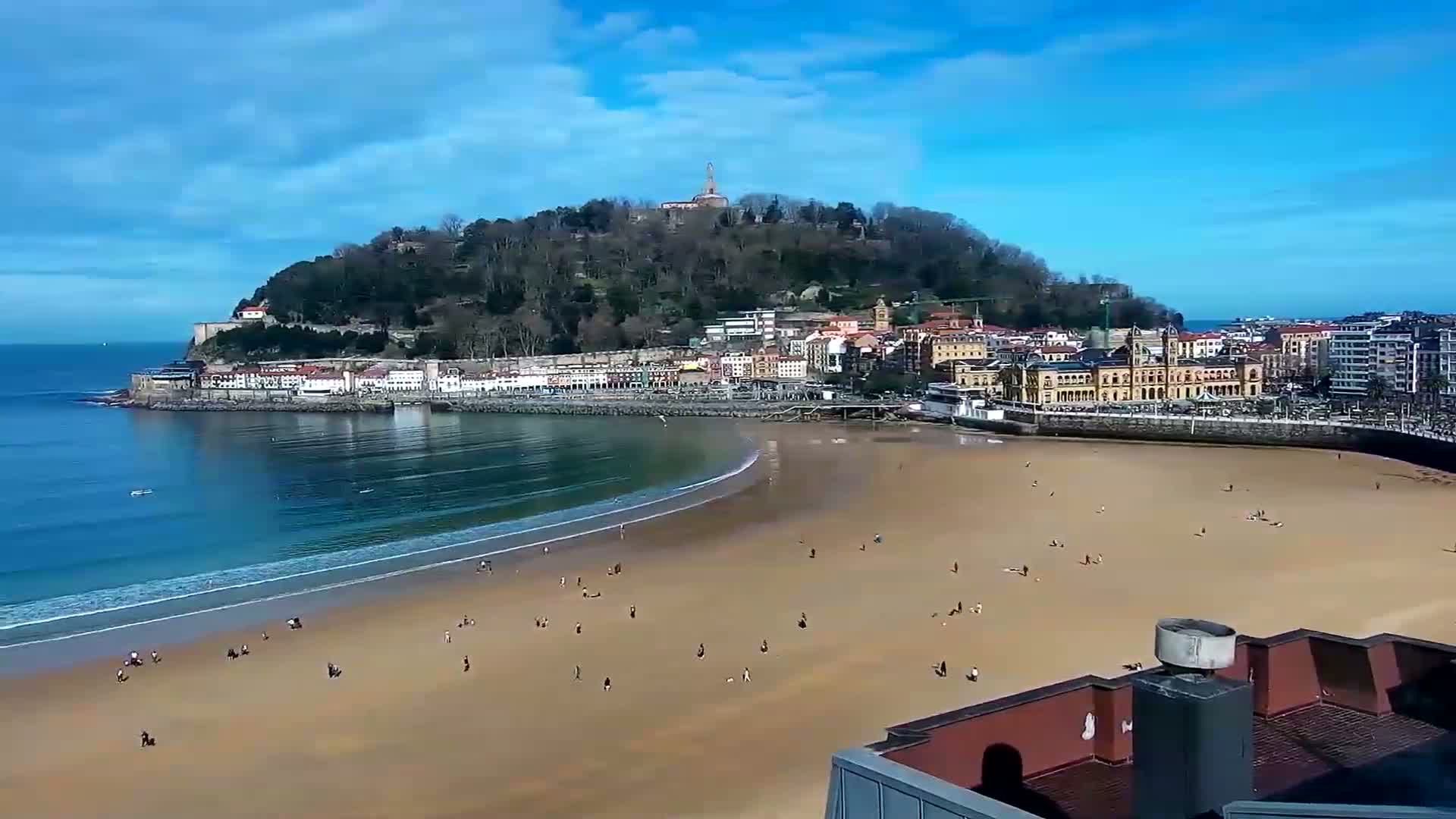A sandy beach with scattered people stretches below a town nestled against a tree-covered hill, all under a mostly sunny sky.