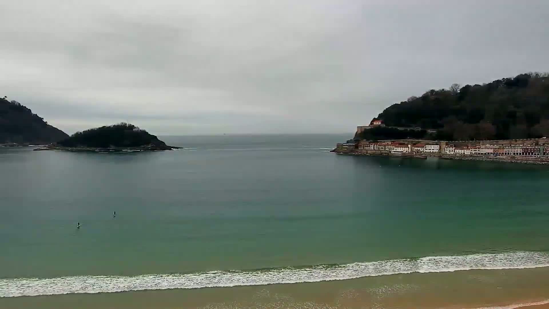 A calm, grey day reveals a coastal scene with two small islands, a town nestled beside a hill, and a sandy beach with gentle waves.