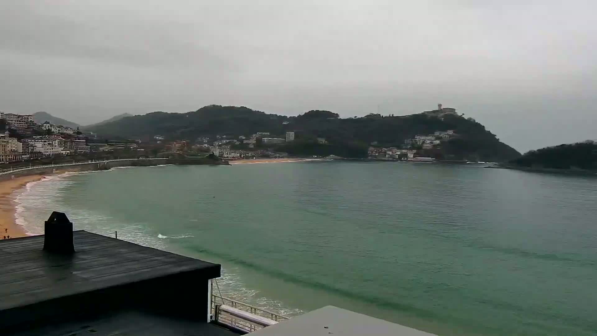 A beach with calm waves curves around a bay under an overcast sky, backed by a town and hills.