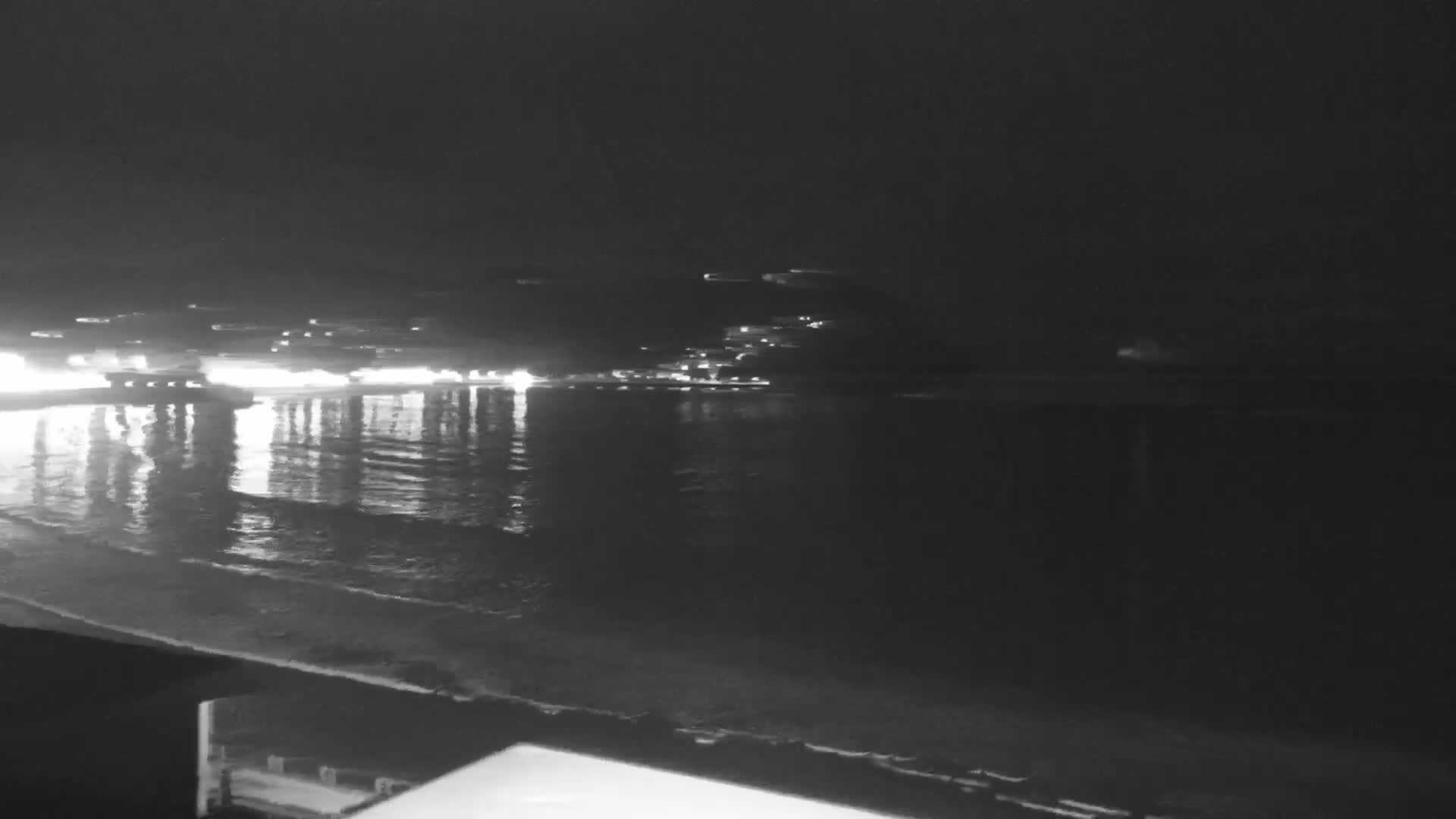 A nighttime view of a calm ocean with lights reflecting on the water from a distant shore.