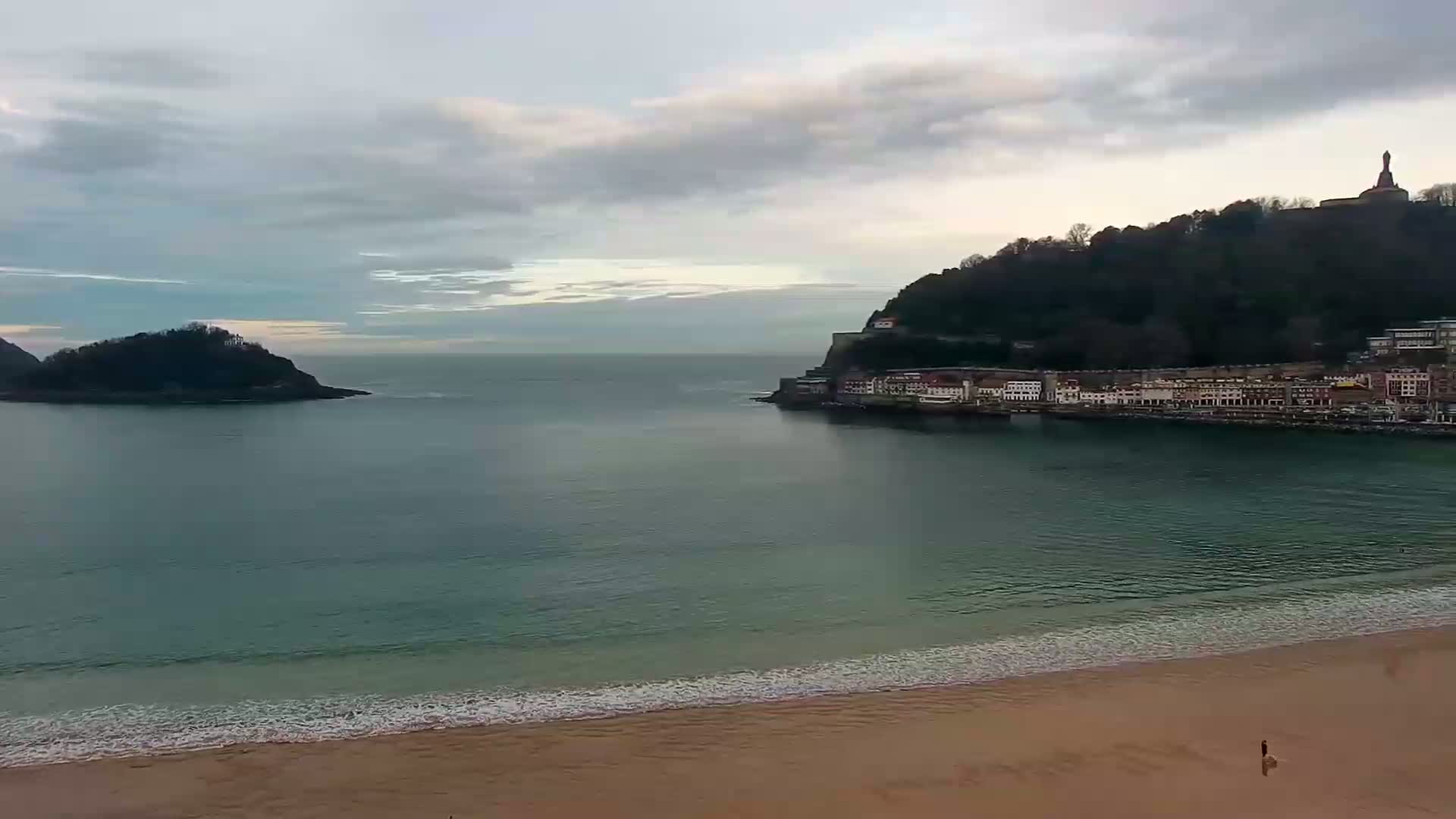 A sandy beach with gentle waves laps the shore, overlooking a calm bay with a small island and a hillside town under a cloudy sky.