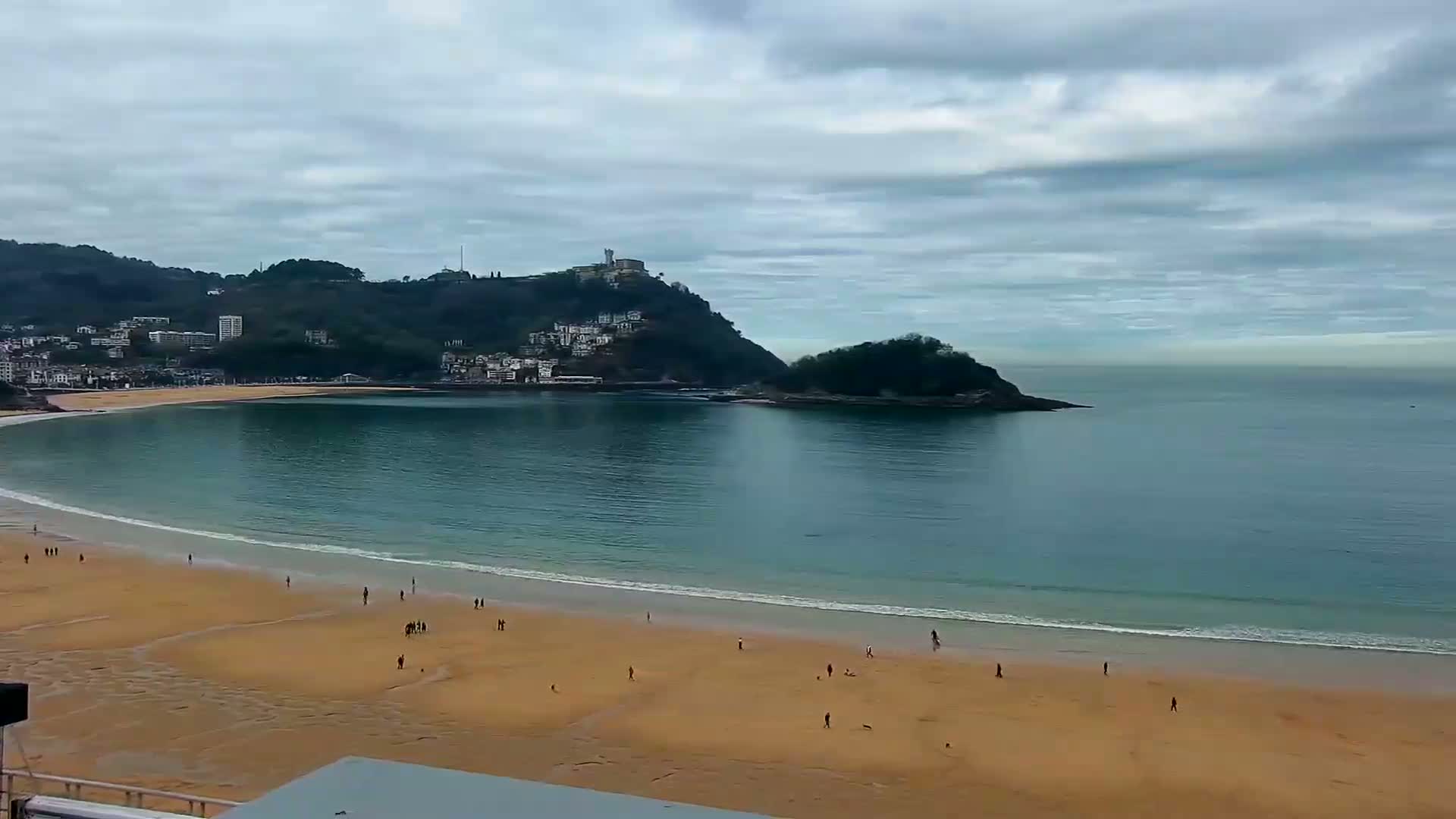 A sandy beach curves around a calm bay under a cloudy sky, with a hill topped with buildings in the background.