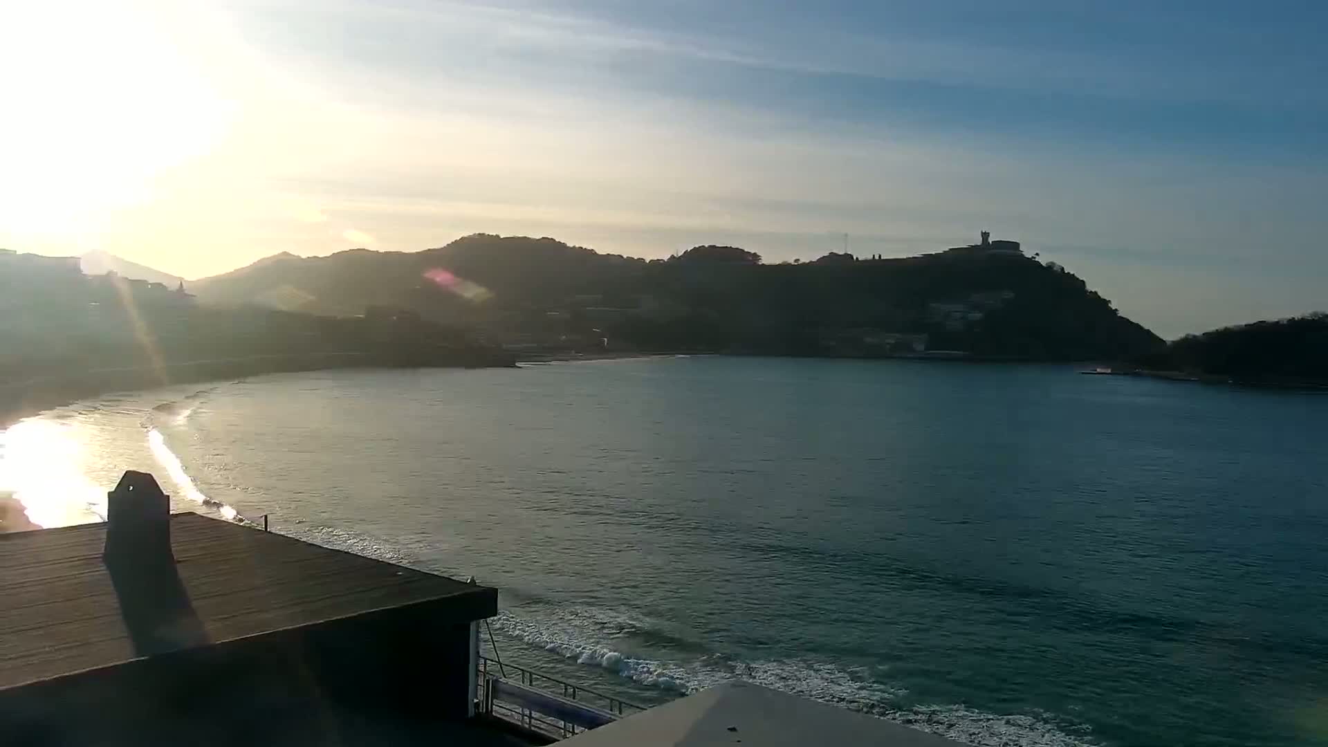 A calm bay with gentle waves laps a sandy beach under a partly sunny sky, hills rise in the background.