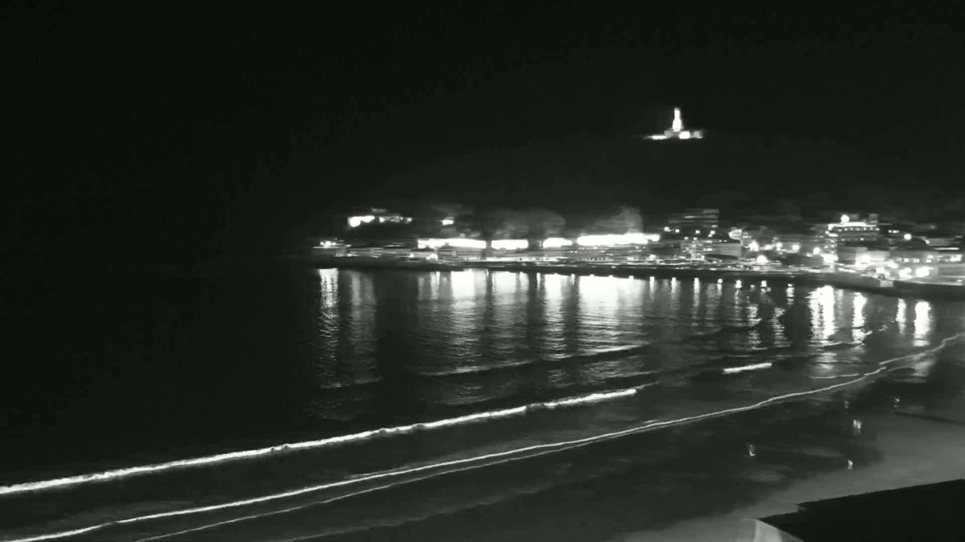 A nighttime, seaside town with gently lapping waves on the shore is illuminated by lights reflecting on the water.