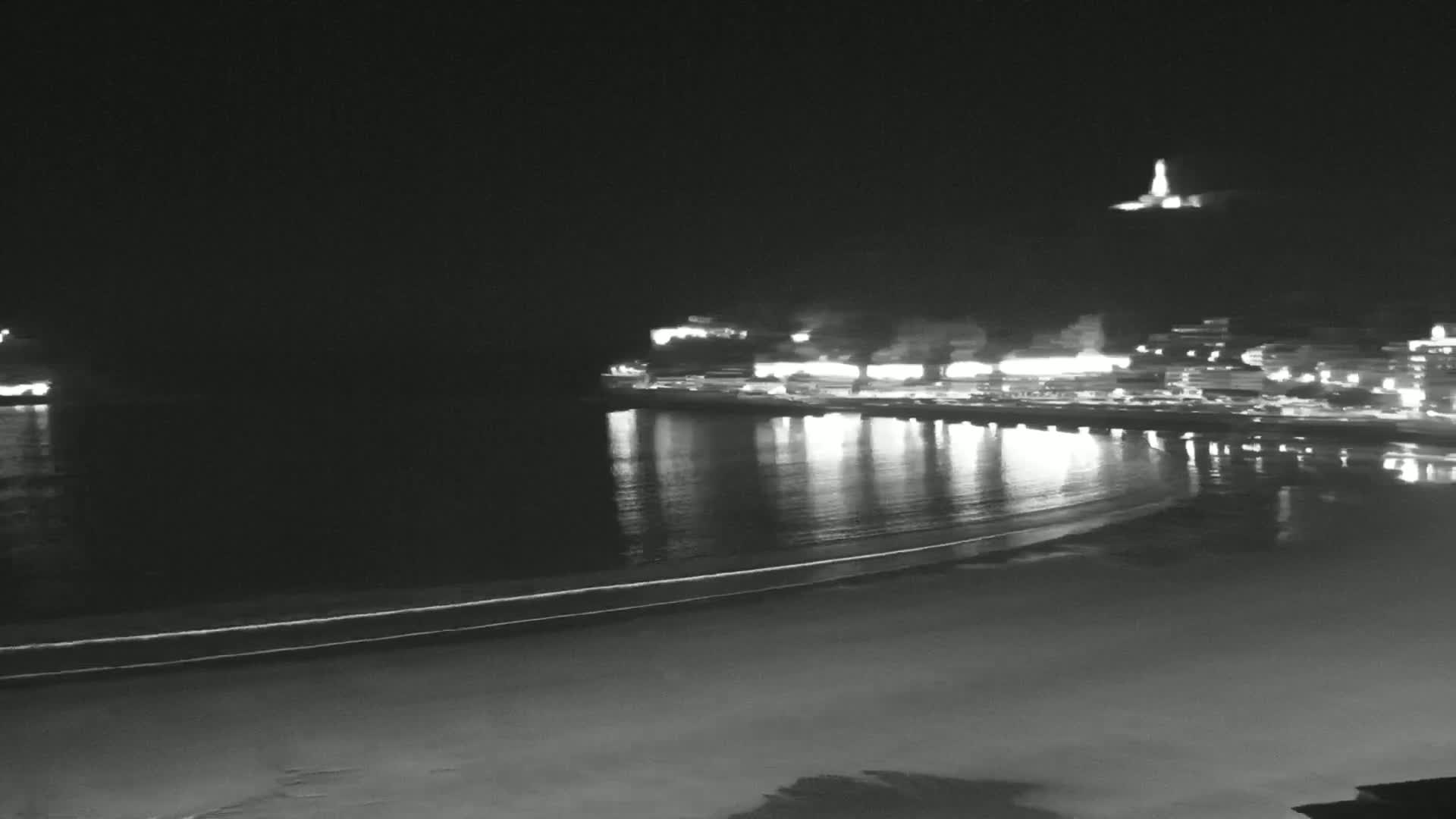 A nighttime, black and white view of a calm, reflective ocean with a brightly lit town on the far shore and a hilltop light in the distance.