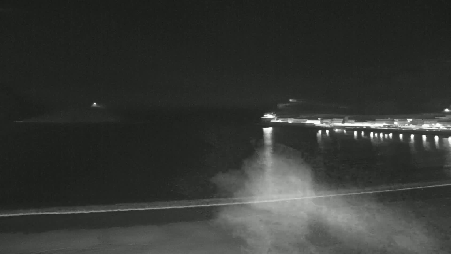 A nighttime, low-light view shows a dark body of water with a shoreline, faint waves, and distant lights on the opposite shore.