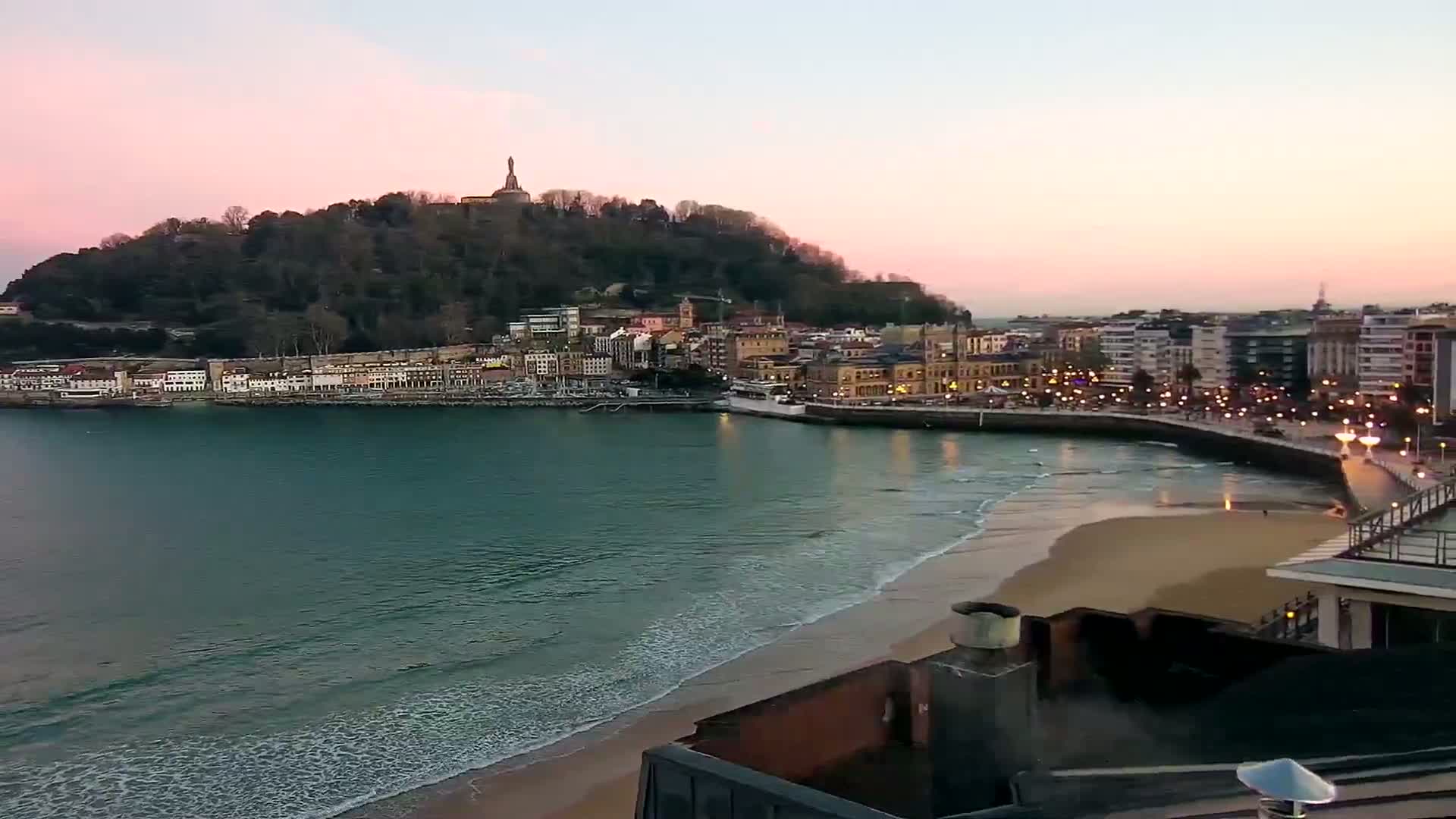 A coastal town at sunset, nestled beneath a hill topped with a monument, features a sandy beach with gentle waves lapping the shore.