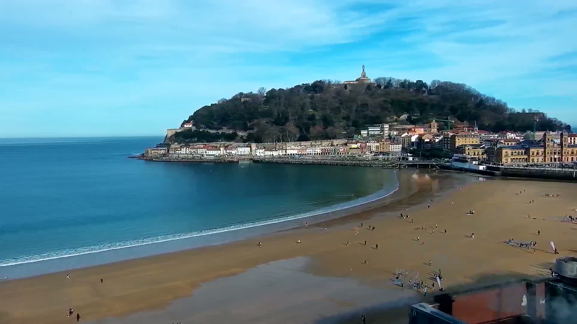 A sandy beach curves around a calm bay, with a town nestled at the base of a hill topped by a monument, all under a clear blue sky.