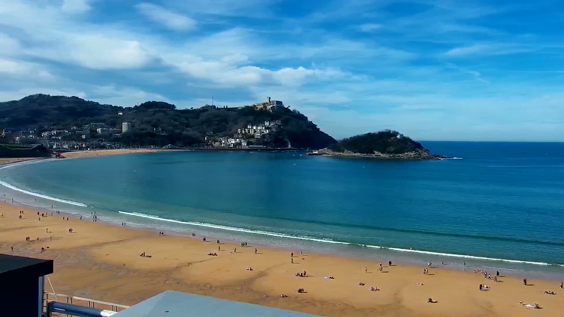 A sandy beach with scattered people curves around a bay, meeting a town nestled at the base of a hill under a mostly sunny sky.