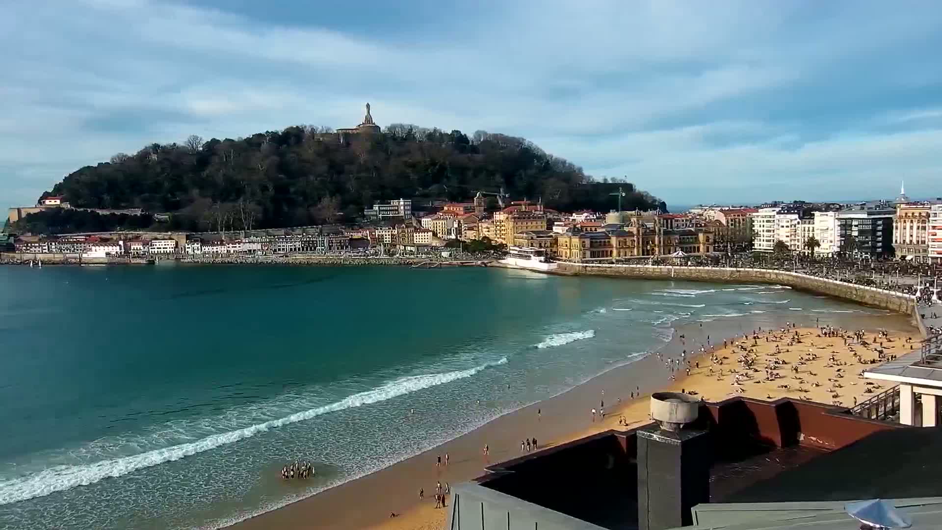 A sandy beach with people sunbathing is bordered by a town nestled at the base of a hill, all under a partly cloudy sky.