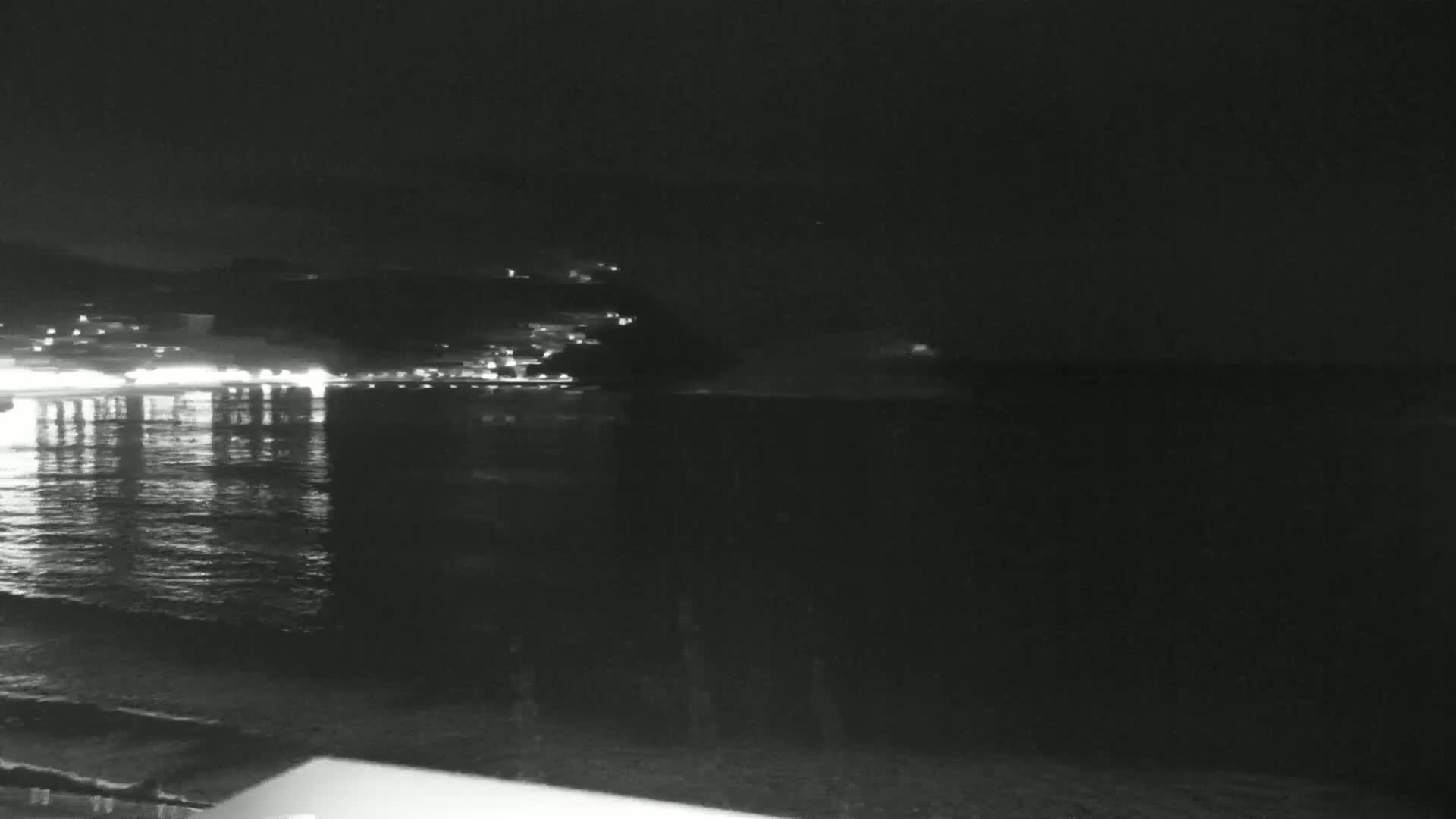 A nighttime coastal scene shows a dark body of water reflecting lights from a distant town on a hill under a clear sky.
